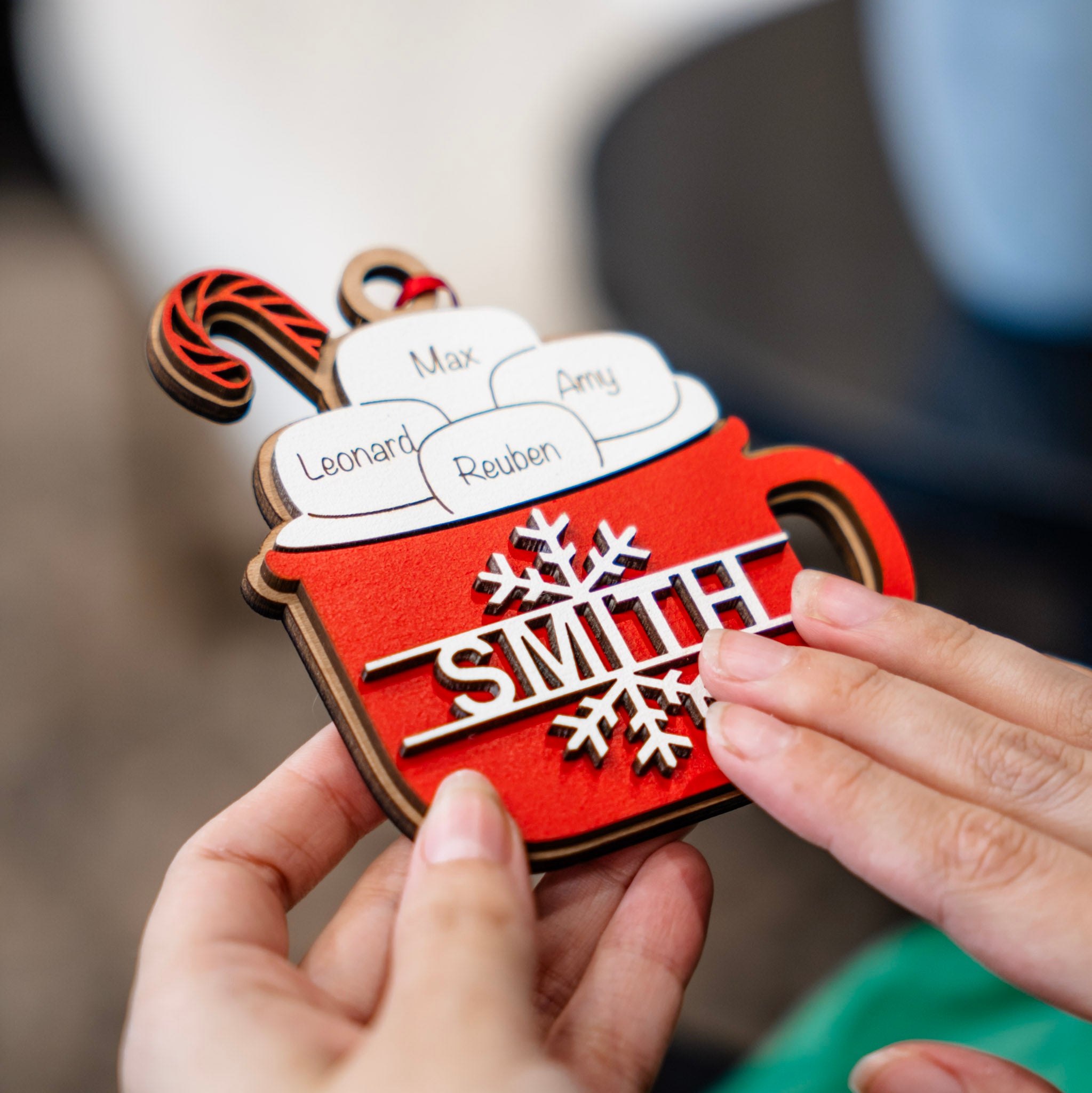 A close-up of a person's hands holding a red and brown wooden Christmas ornament shaped like a hot cocoa mug. The ornament features white foam swirls with names printed on them (Max, Leonard, Reuben, Amy), a red-and-brown striped candy cane, and a silver metal nameplate that reads 'SMITH' with snowflake details.