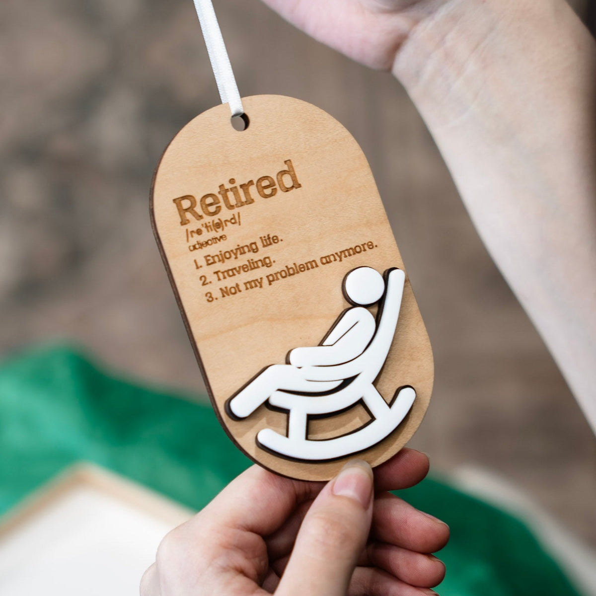 A person holds a wooden ornament with a white ribbon. The tag is engraved with the word 'Retired' and a definition, with a white silhouette of a person sitting in a rocking chair.