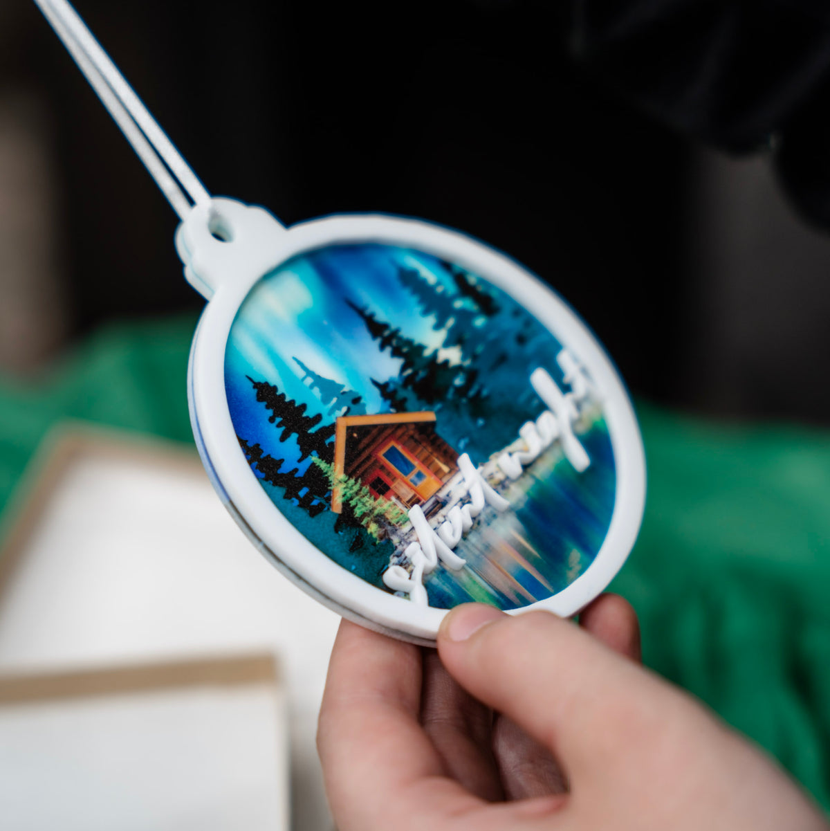 A hand holds a round white ornament featuring a painted cabin scene by a blue lake with pine trees and the words 'silent night' in white script. A green fabric backdrop and part of a gift box are visible in the background.