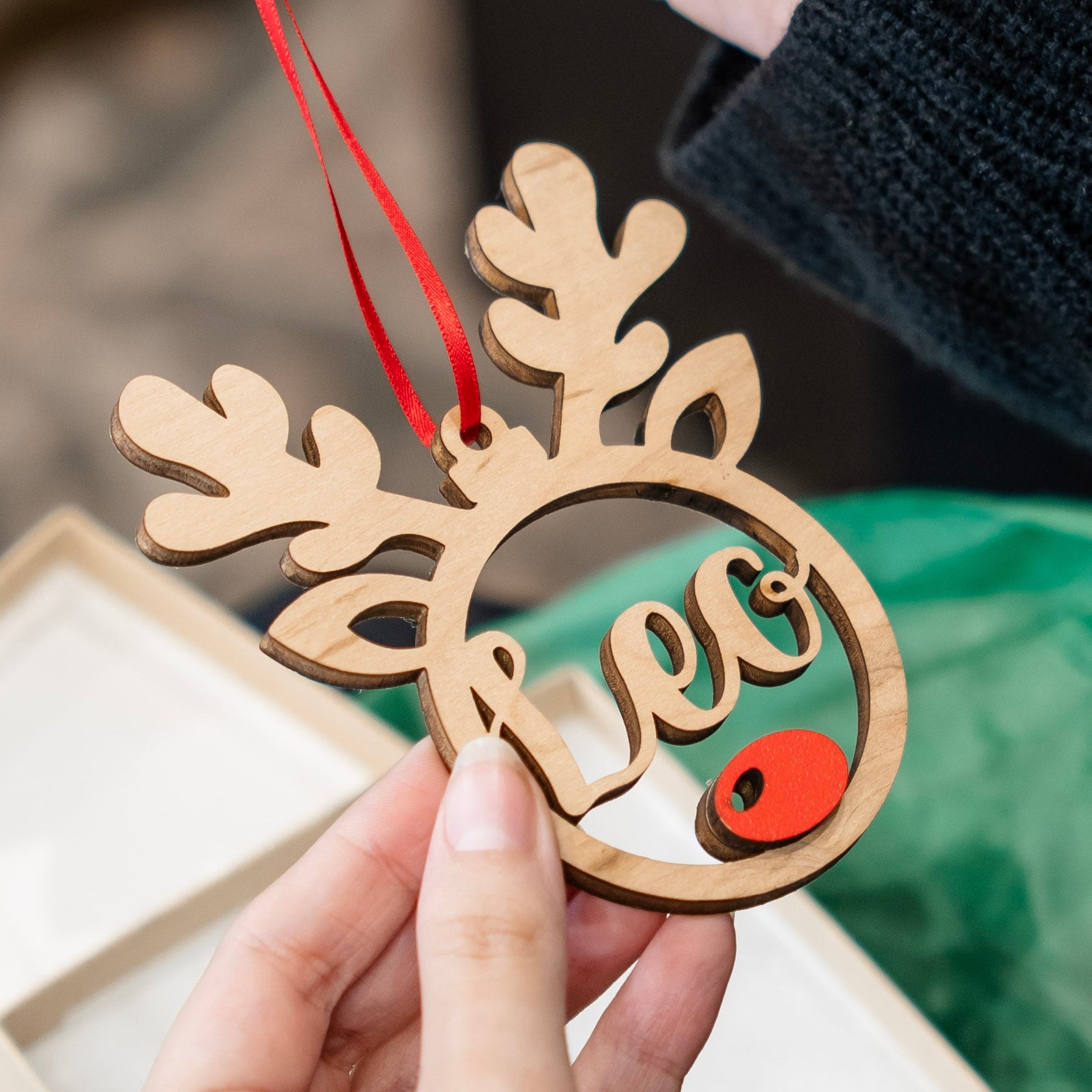 A wooden reindeer-shaped Christmas ornament with a red hanging ribbon, held by hands over a gift box. The ornament features a laser-cut reindeer head silhouette with the name 'Leo' in cursive across the face and a small red wooden nose.