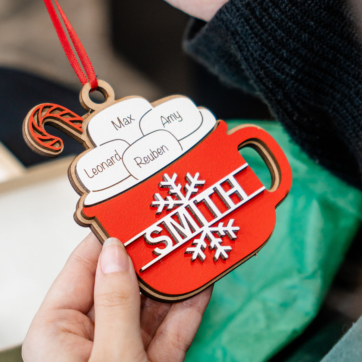 A hand holds a personalized wooden Christmas ornament shaped like a red hot cocoa mug with white foam and the name 'SMITH' in silver, along with custom names on the foam and a red hanging ribbon.