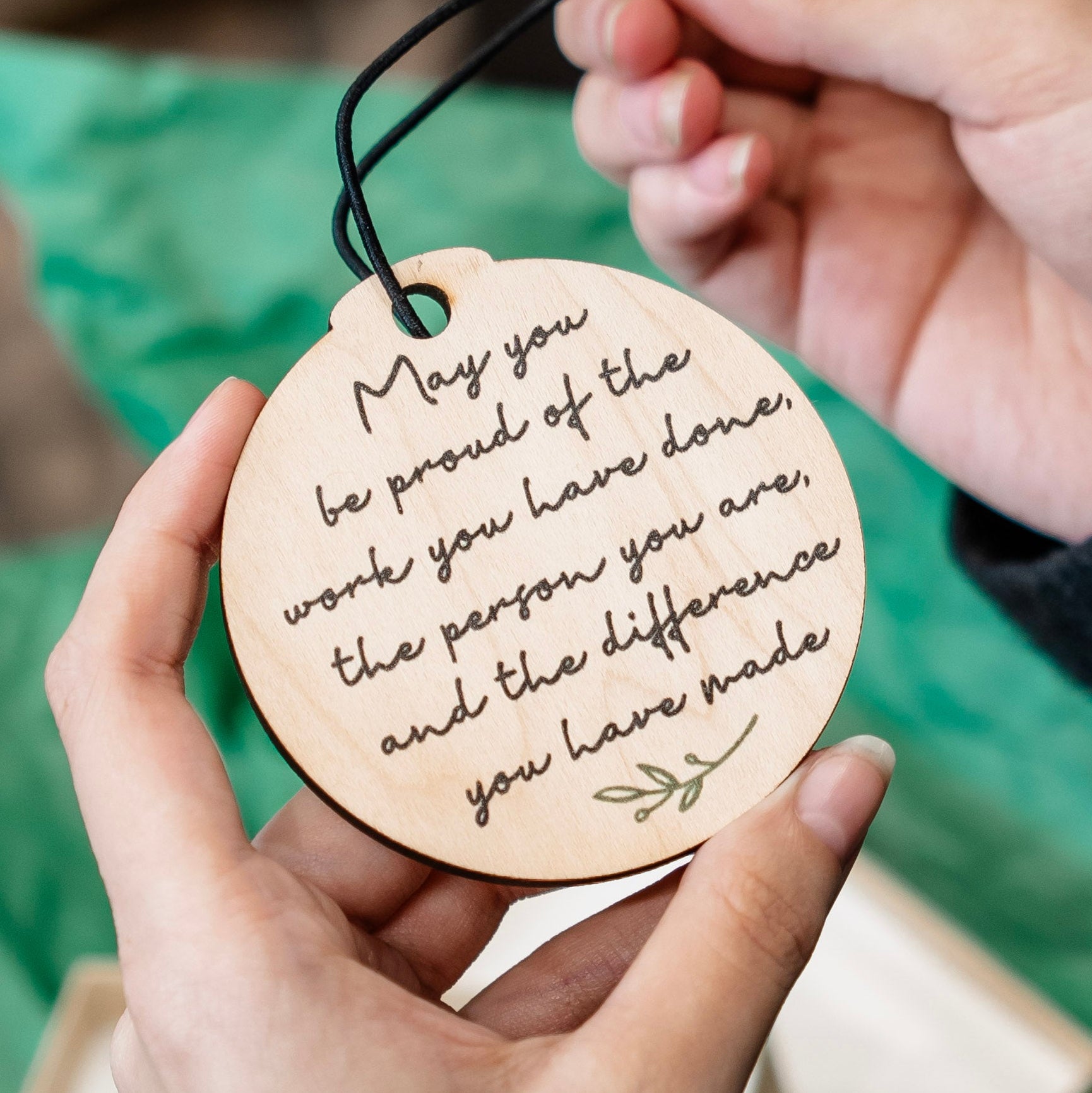 A person holds a round wooden ornament with a black hanging stretchy string. The ornament is engraved with a motivational quote and a small leaf illustration.