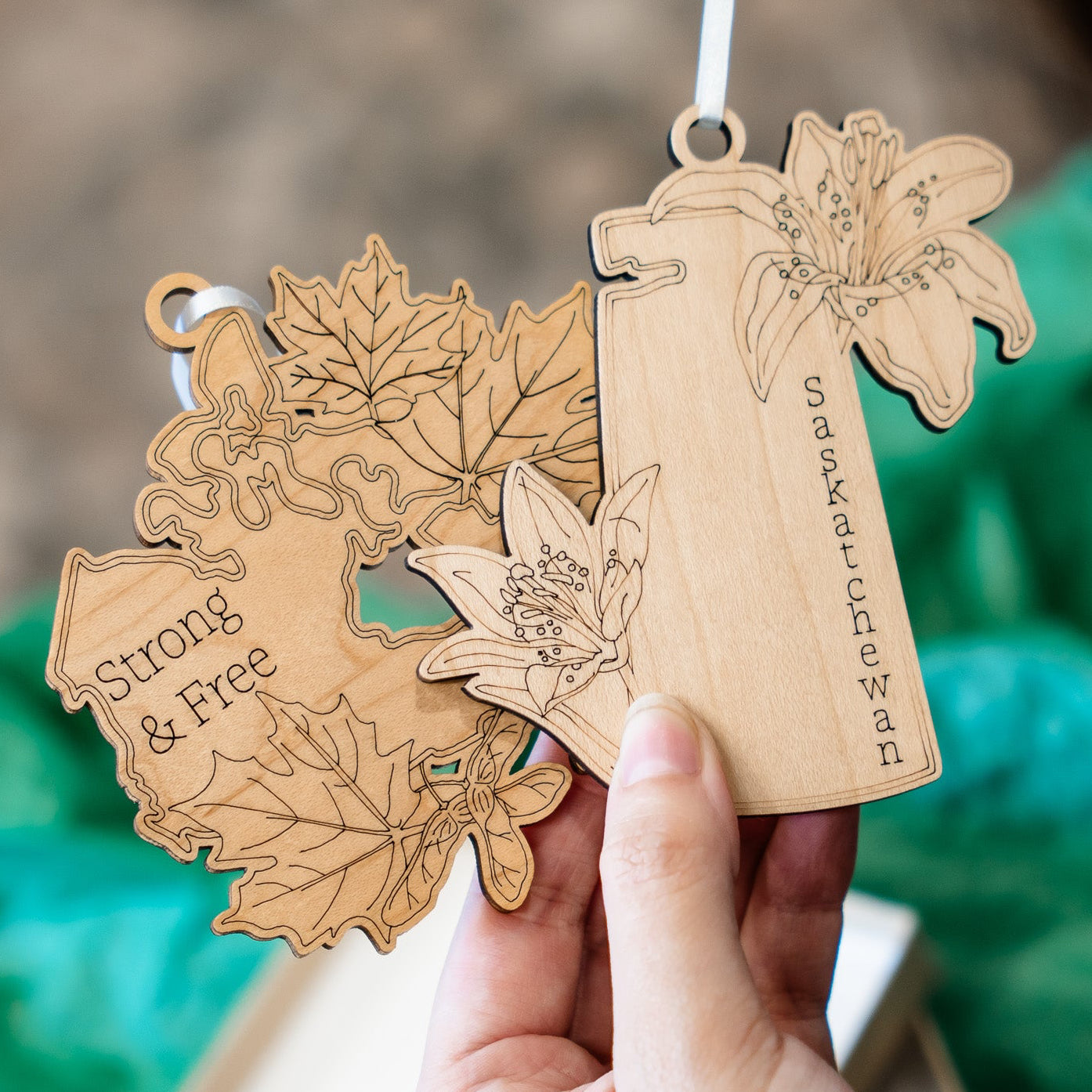 A hand holds a wooden ornament featuring engraved floral and puzzle-piece designs with the words 'Strong & Free' and 'We are all just trees.' The background is softly blurred with green tissue paper and a neutral surface.