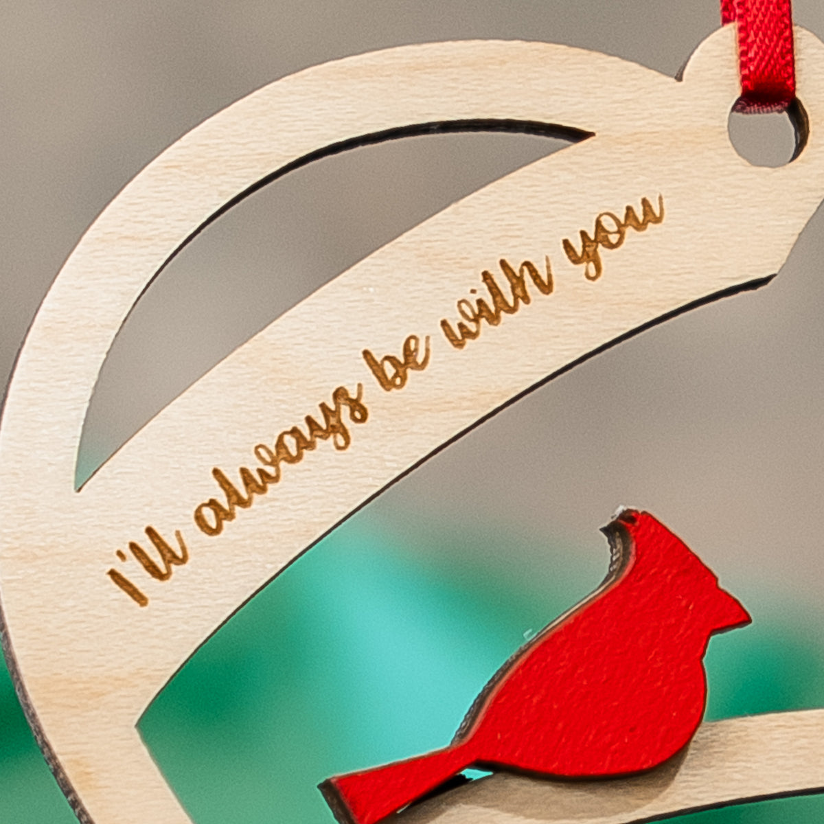 A close-up of a wooden Christmas ornament featuring a red cardinal and the engraved phrase 'I'll always be with you' in gold-toned lettering, with a red hanging ribbon visible at the top.