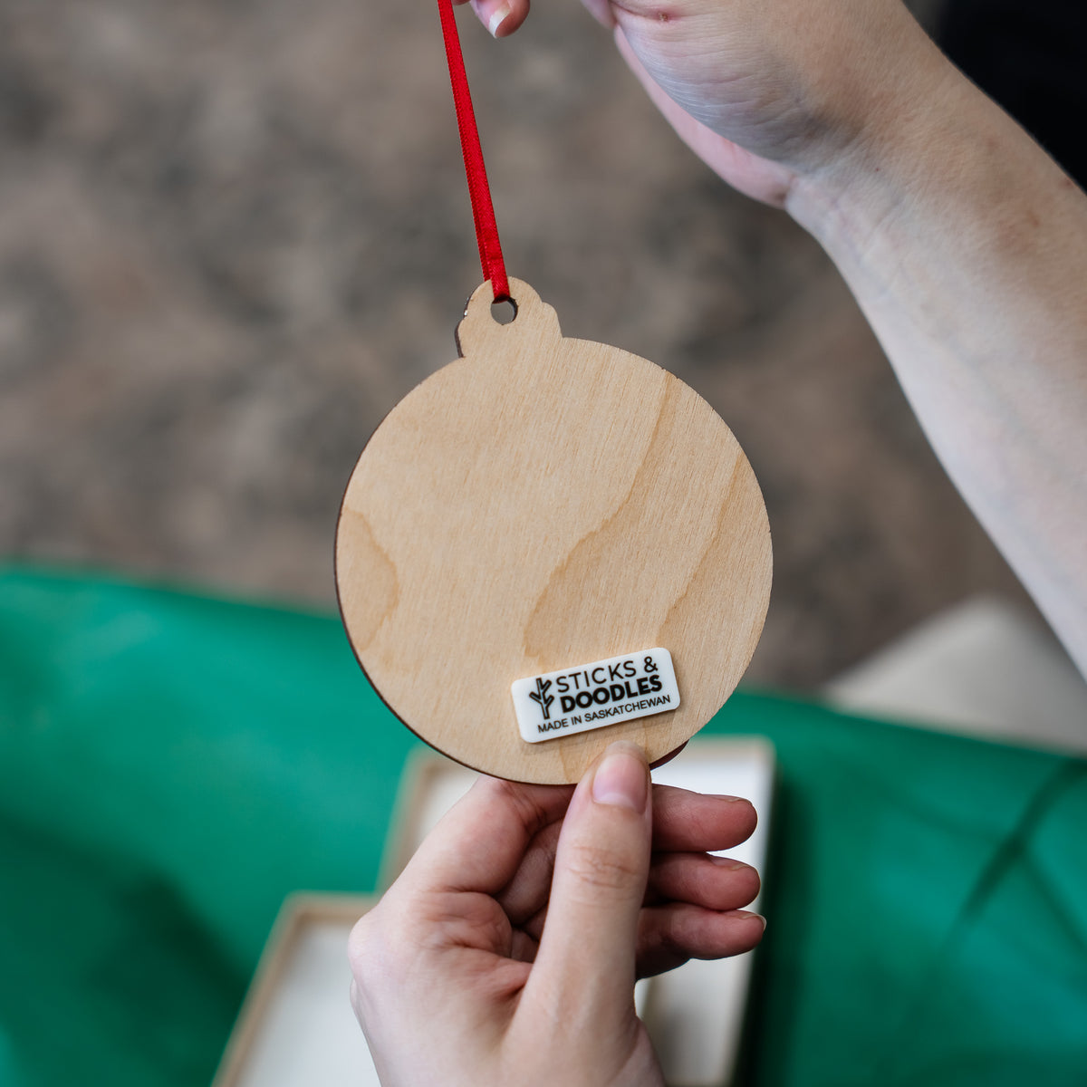 A person holds a round natural wood ornament with a red hanging string. The ornament has a small white sticker that reads 'STICKS & DOODLES MADE IN Saskatchewan'.