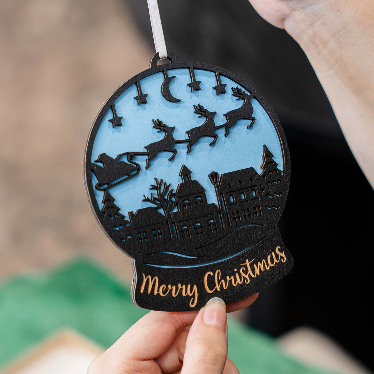 A hand holds a round black Christmas ornament featuring a snow globe design with Santa, sleigh, stars, moon, houses, and the words 'Merry Christmas' in gold, against a softly blurred background.
