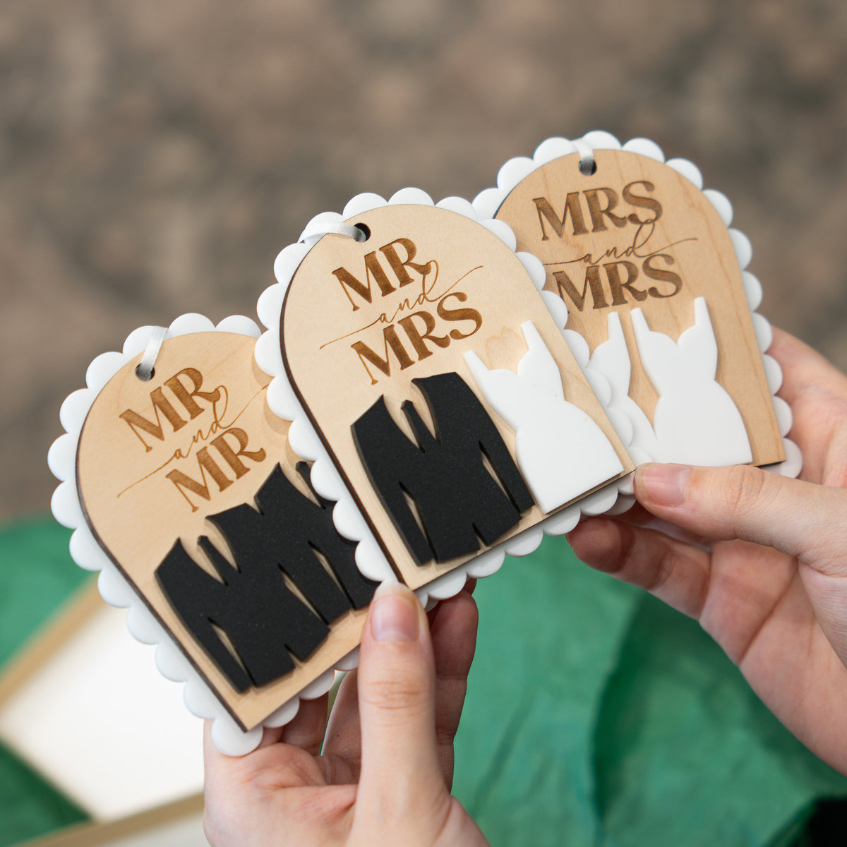 Three scalloped wooden gift tags with laser-engraved 'MR and MRS' designs are held in two hands. The tags feature different color combinations: one black and tan, one black and white, and one white and tan, with a blurred green and brown background.