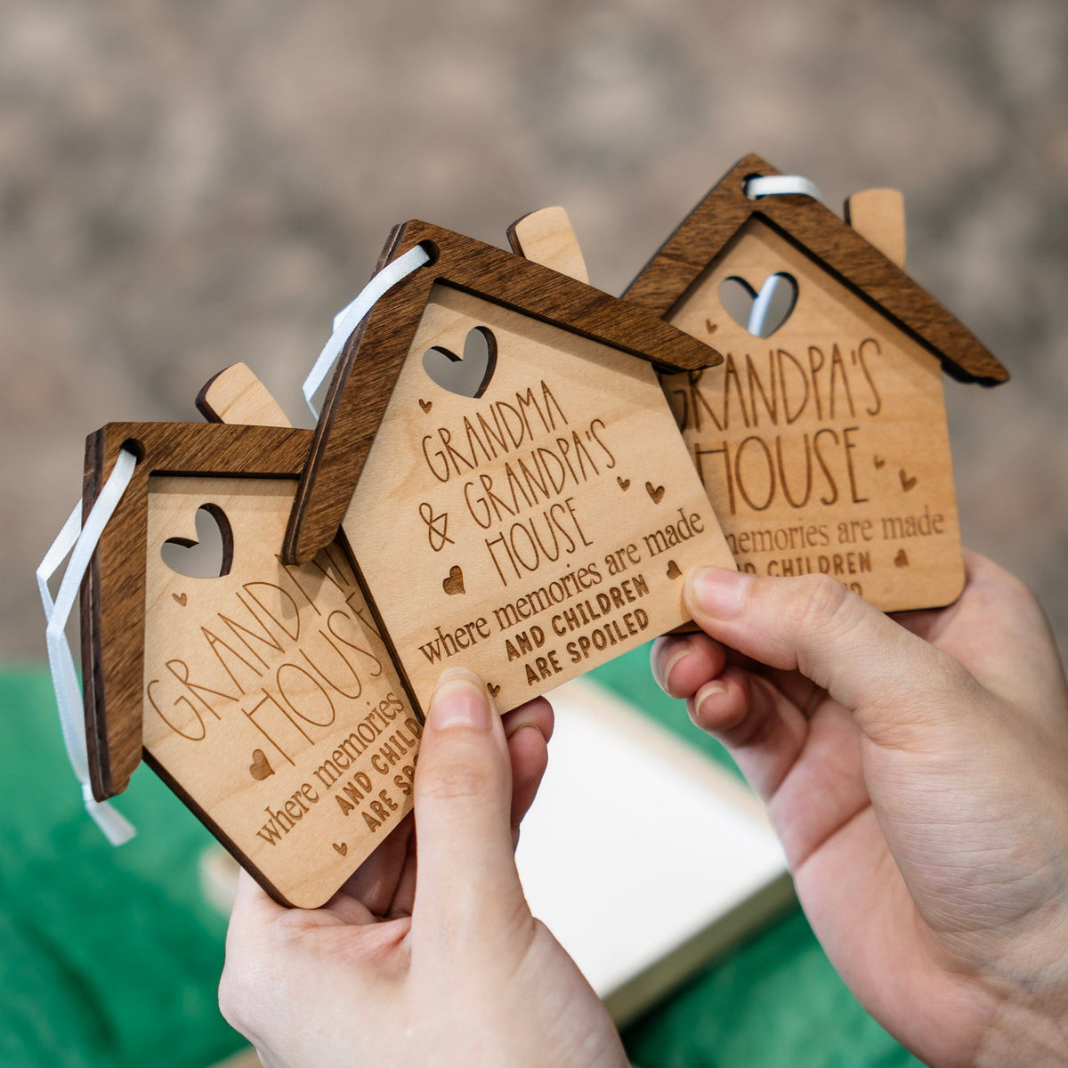 Three personalized wooden house-shaped ornaments being held by hands, engraved with 'GRANDMA & GRANDPA'S HOUSE' and small heart cutouts, against a softly blurred outdoor background.
