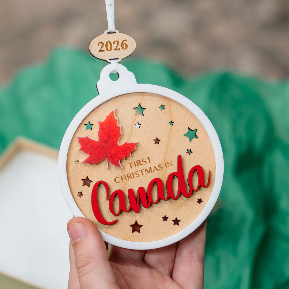 A hand holds a round wooden Christmas ornament engraved with '2026', 'FIRST CHRISTMAS IN Canada', a red maple leaf, and small star cutouts. The ornament is shown against blurred green tissue paper background with part of a gift box visible.