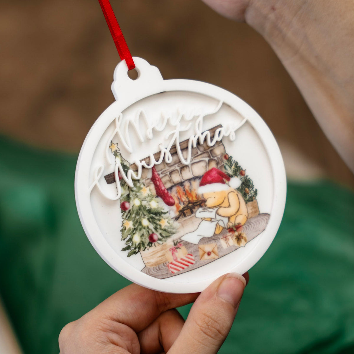 A person holds a round white Christmas ornament with a red ribbon. The ornament features a detailed holiday illustration of a Pooh bear around a cozy fireplace scene with a tree, gift boxes, and the words 'Merry Christmas' in white script.