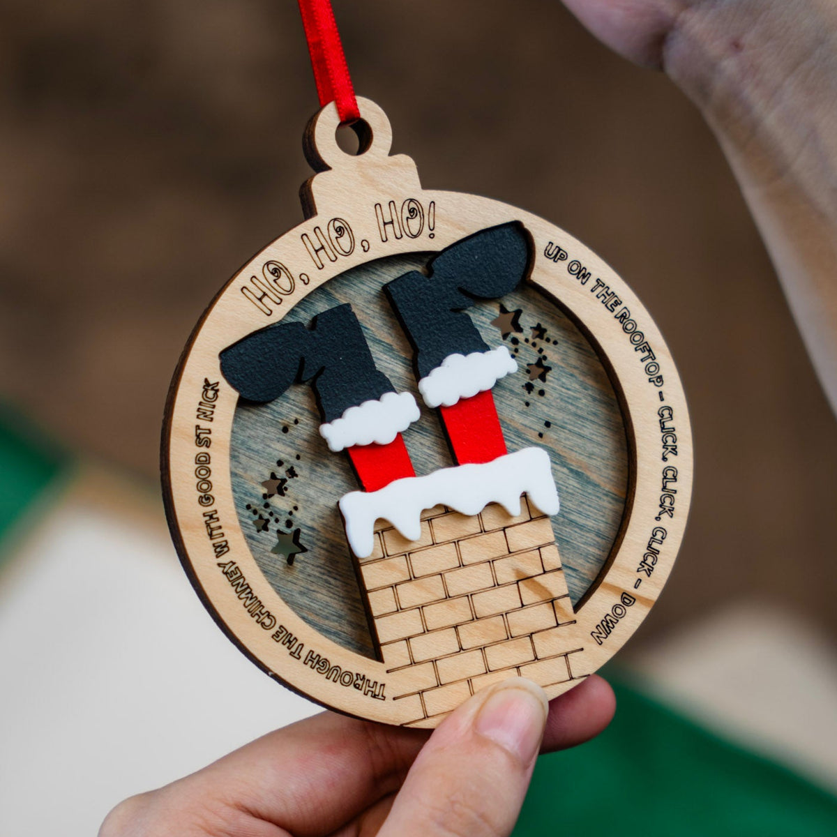A close-up of a hand holding a personalized round wooden Christmas ornament, with Santa's feet upside down, stuck in a chimney. The ornament features a laser-engraved background with the words 'HO HO HO!' and the lyrics from Up on a house top.