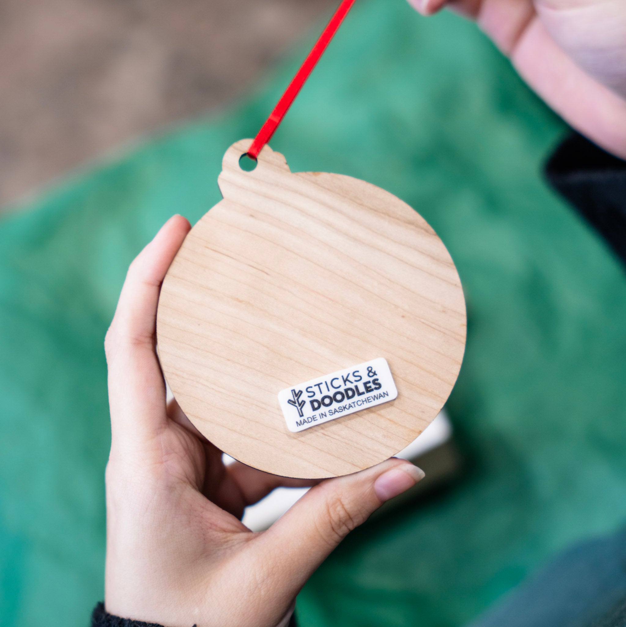 A hand holds a round wooden dog treat puzzle with a red hanging string and a white 'STICKS & DOODLES' label. The background is a soft-focus green surface.
