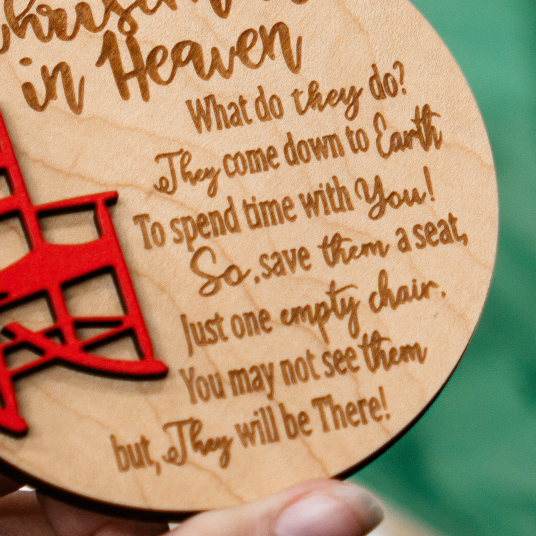 A close-up of a round wooden plaque held in a hand, featuring engraved text about heaven and a red laser-cut chair design on a green background.