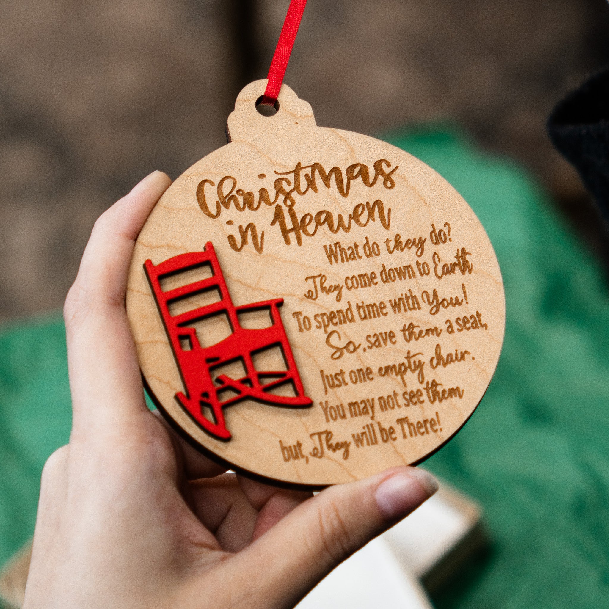 A hand holds a round wooden Christmas ornament with a red hanging ribbon. The ornament is engraved with the words 'Christmas in Heaven' and a poem, with a red illustration of a ladder leading up toward a Christmas tree.