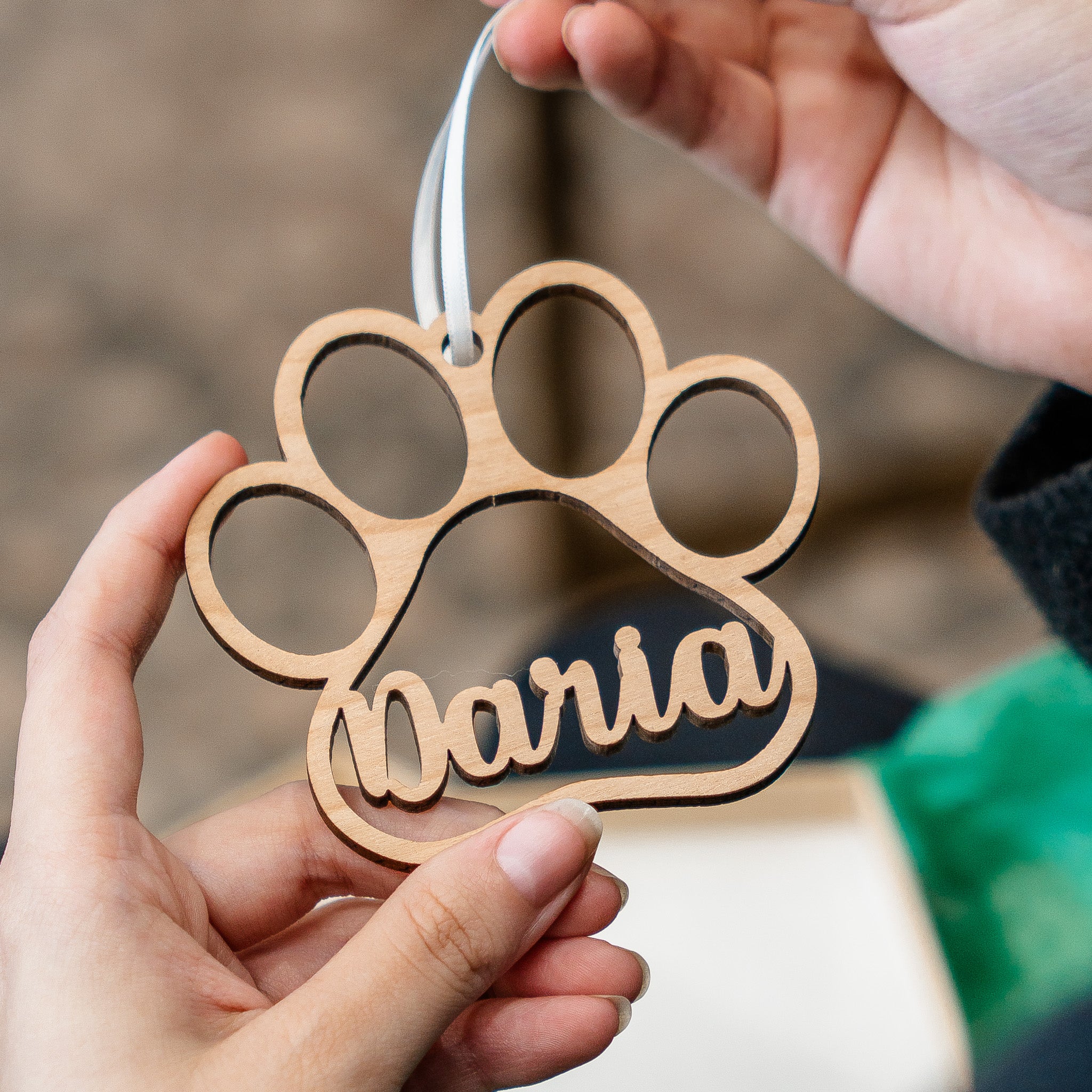 A wooden paw-shaped ornament with the name 'Naria' cut into the pad, held by two hands against a softly blurred outdoor background.