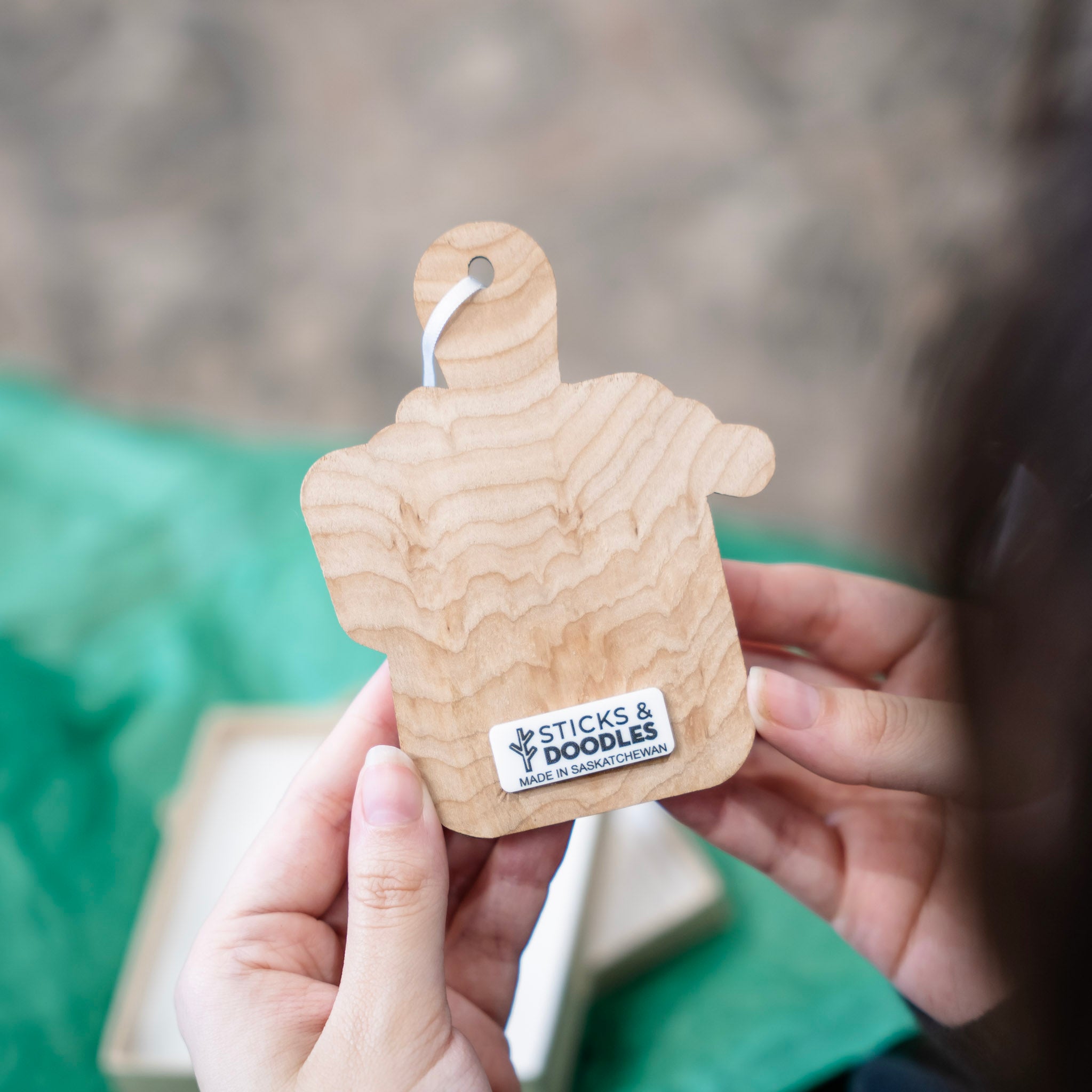 A person holds a small wooden baking tray shaped ornament with a visible wood grain pattern and a white sticker that reads 'STICKS & DOODLES MADE IN SASKATCHEWAN'. The background is heavily blurred with green fabric and a neutral surface.