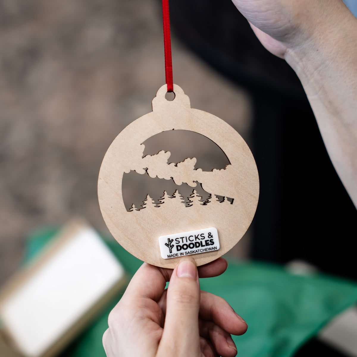 The back of a personalized laser-engraved ornament held by hands, with a red ribbon, and a tag with 'Sticks & Doodles Made In Saskatchewan'.