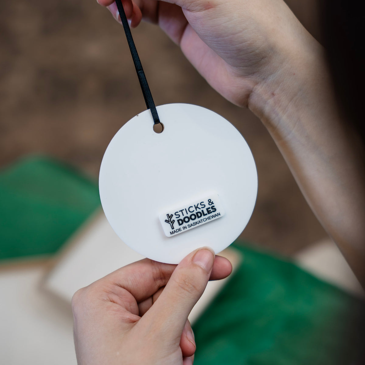 A person holds the back of the ornament from a black ribbon that is white acrylic with a tag that says Sticks & Doodles Made in Saskatchewan. 