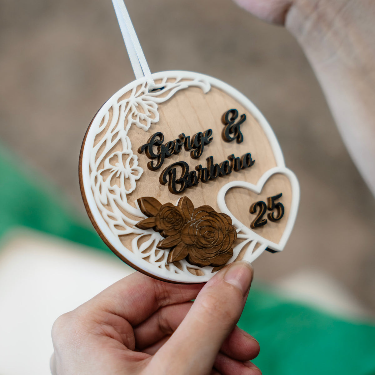 A hand holds a personalized round wooden ornament with laser-cut floral details, the names 'George & Barbara,' and the number '25' in dark brown, against a softly blurred outdoor background.