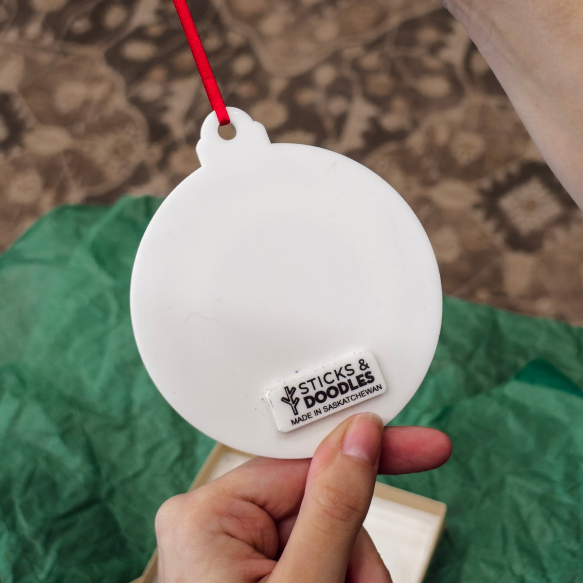 Back of ornament made with white acrylic and hanging with a red ribbon, with a tag that says Sticks & Doodles Made in Saskatchewan.