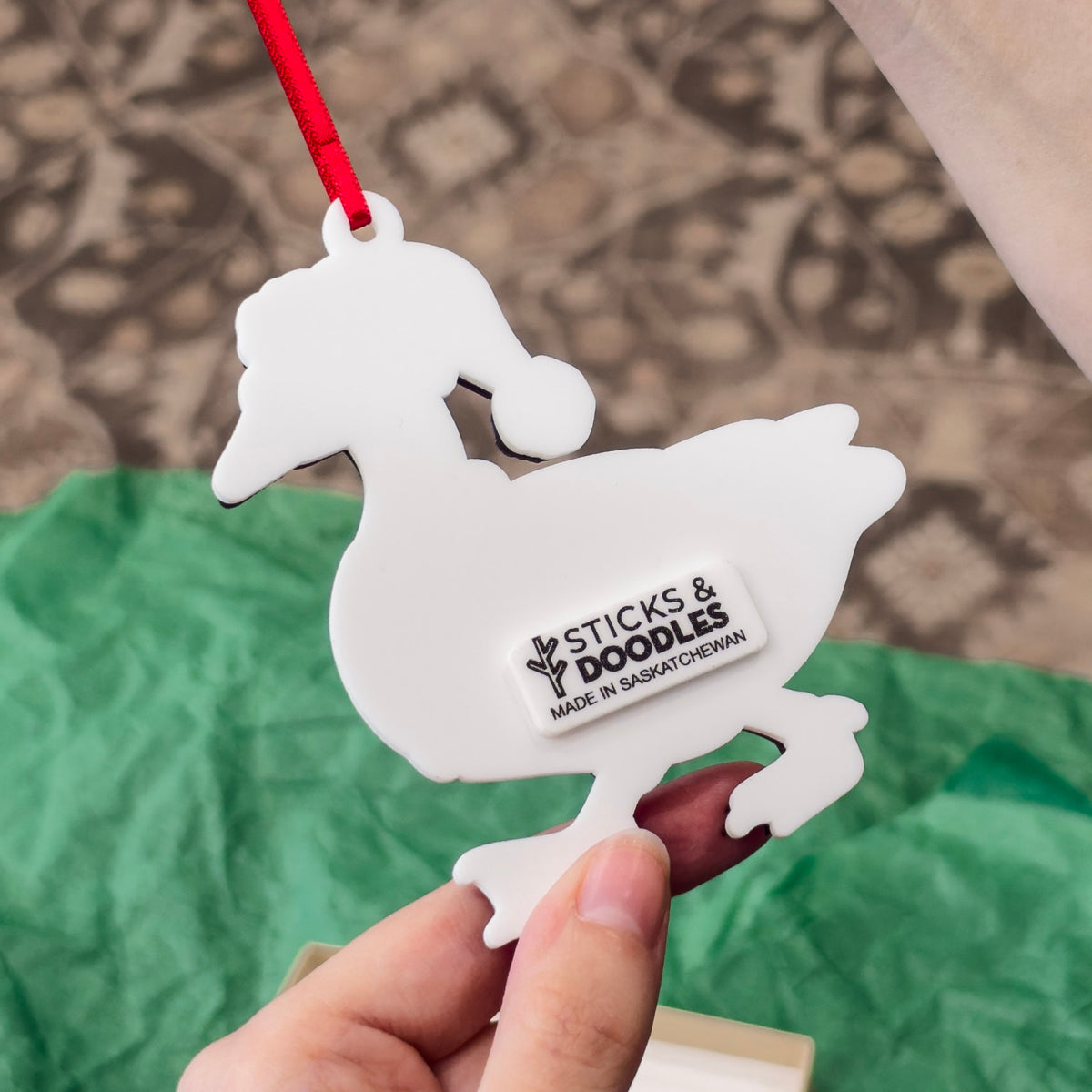 Back of ornament made with white acrylic, shaped like a goose wearing a santa hat, with a tag that has Sticks & Doodles Made in Saskatchewan tag on the back.