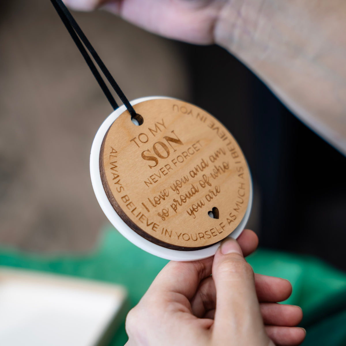 Hands hold a round wooden ornament with a black hanging ribbon. The ornament is engraved with the message 'TO MY SON NEVER FORGET I love you and am so proud of who you are' and features a white outer layer.
