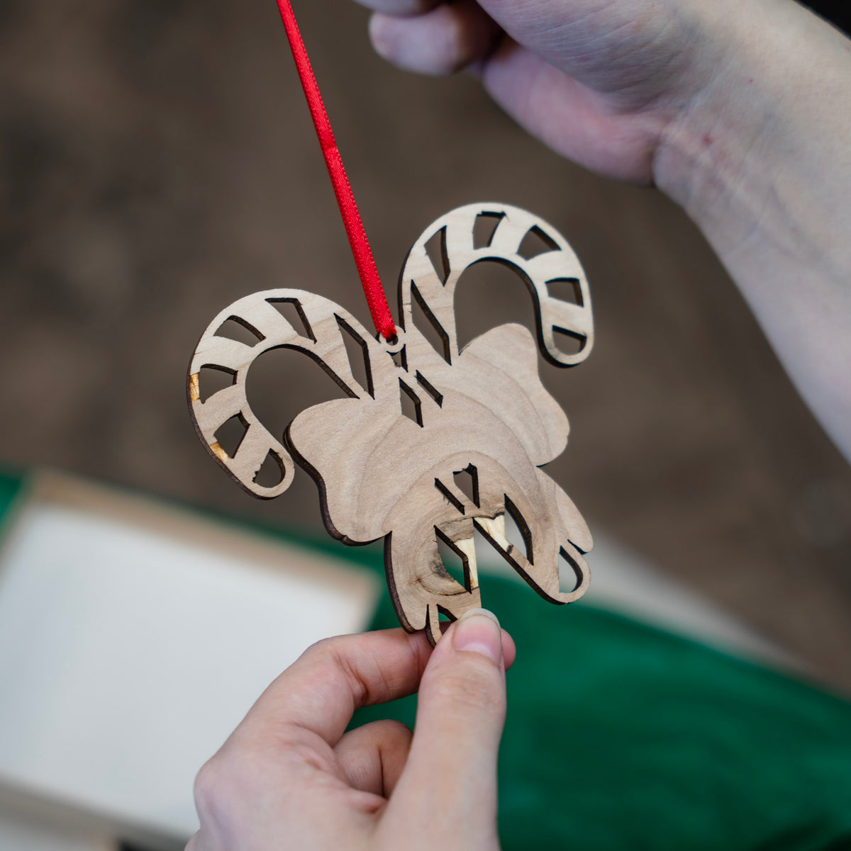 A close-up of hands holding the backside of the laser-cut wooden Christmas ornament shaped like two candy canes with a red bow, suspended by a red ribbon against a soft-focus background of green tissue paper and a gift box.