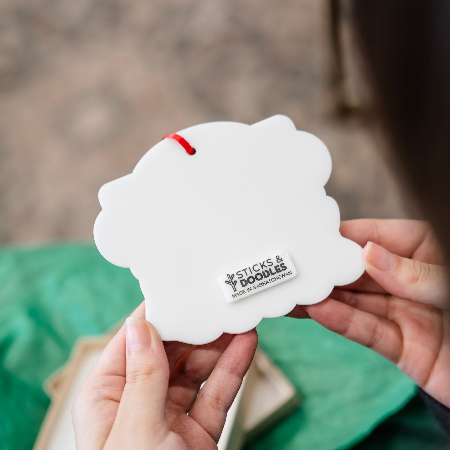 A person holds a white acrylic backer of an ornament with a tag that says: 'STICKS & DOODLES, SASKATCHEWAN', featuring a small red ribbon.