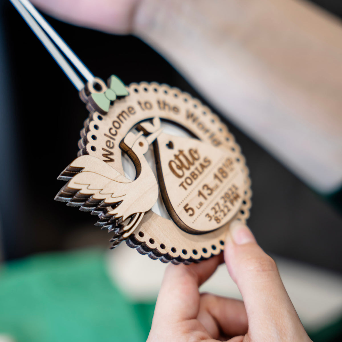 A hand holds a personalized wooden baby shower ornament shaped like a stork with a green bow, hung with a white ribbon, featuring engraved text reading 'Welcome to the World' and 'Otto Tobias' with birth details. The background is softly blurred with green fabric and neutral tones.
