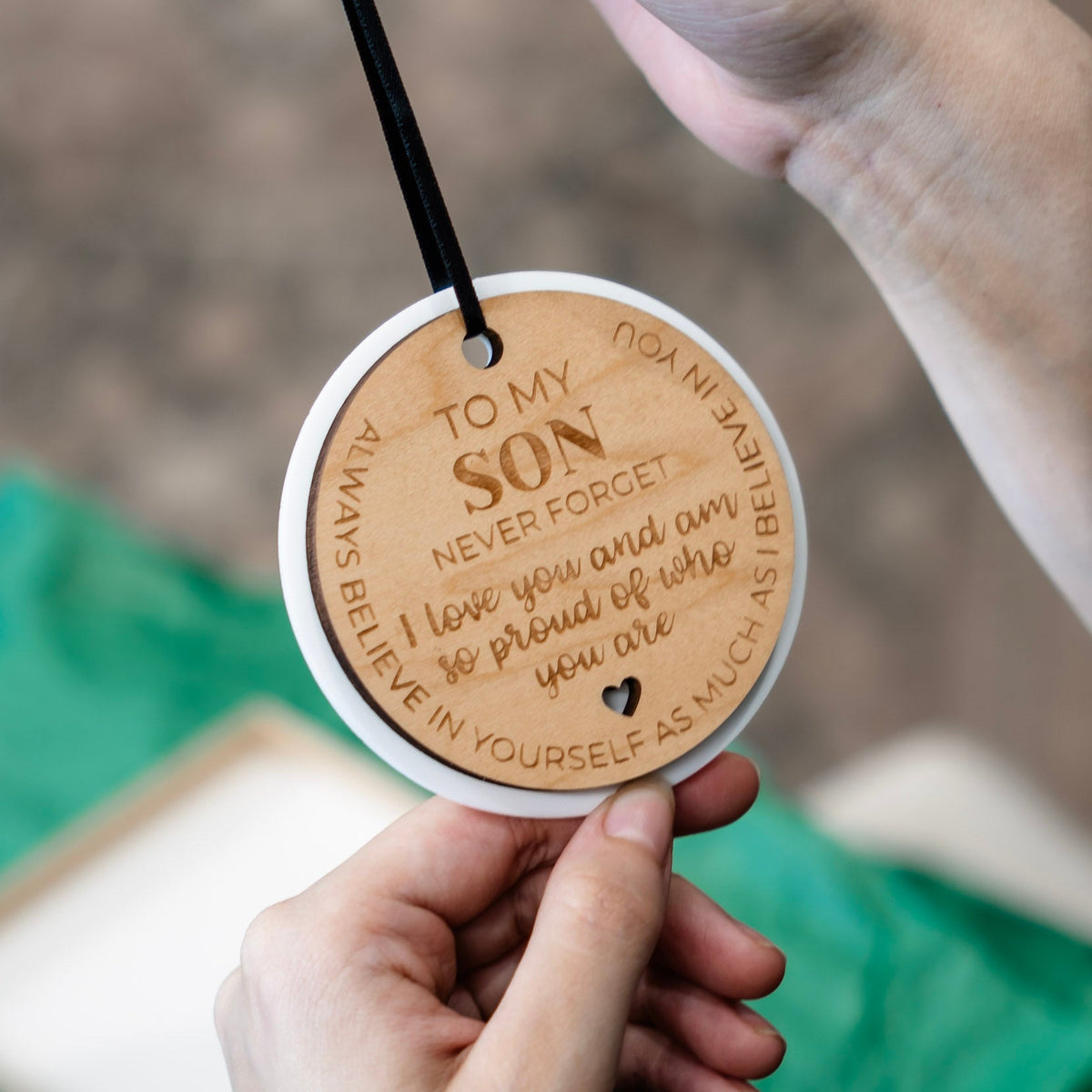 Hands hold a round wooden ornament with a black hanging ribbon. The ornament is engraved with the message 'TO MY SON NEVER FORGET I love you and am so proud of who you are' and features a white outer layer.