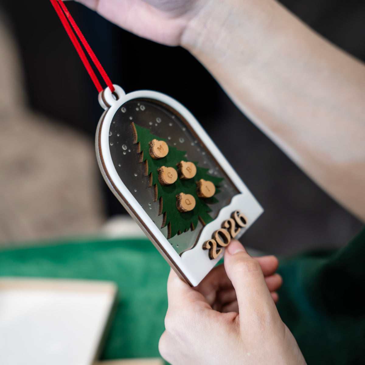 A person holds a personalized wooden Christmas ornament shaped like a tree with the year '2026' and small wooden ornaments for initials, hanging it by a red ribbon. The background is softly blurred with a green fabric surface.