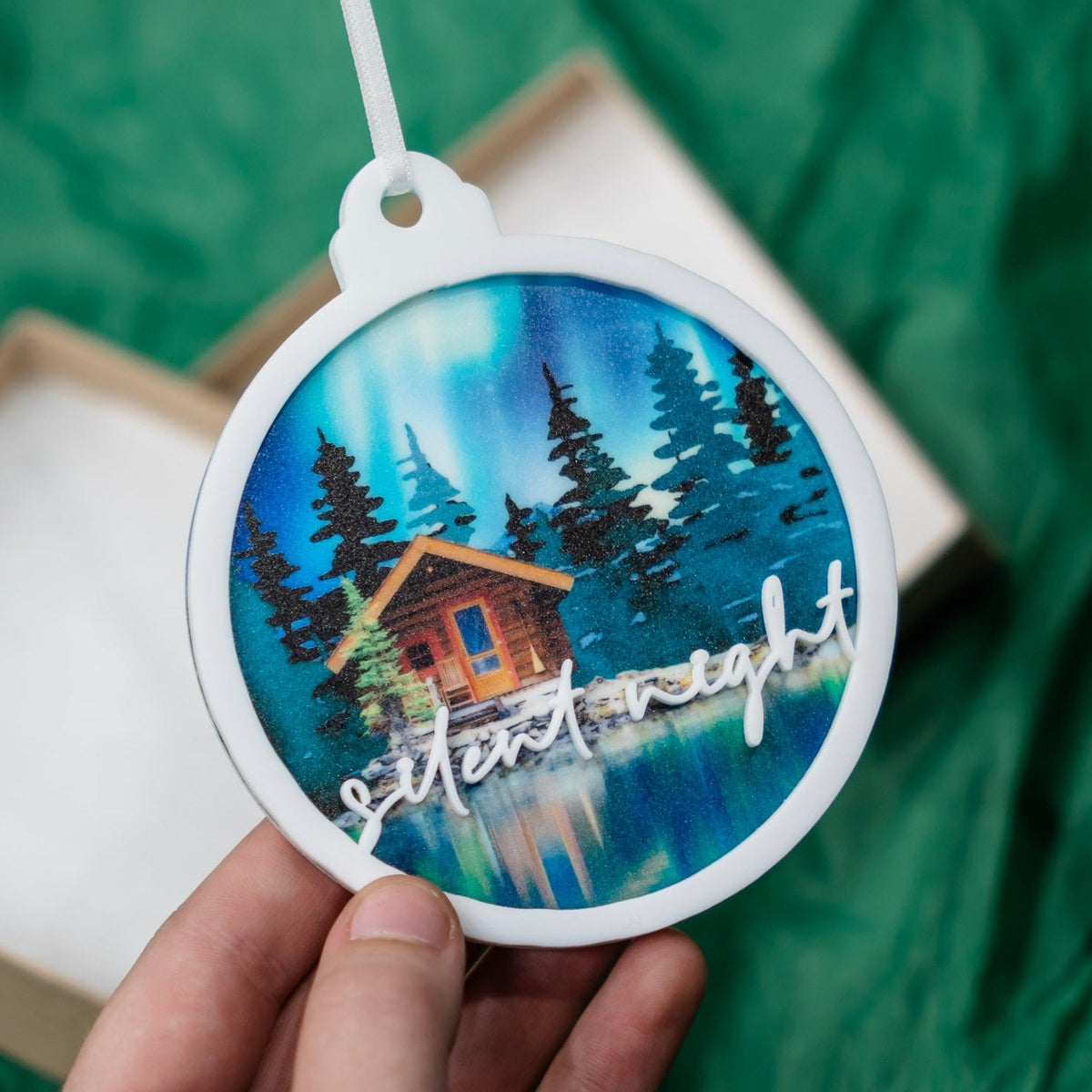 A hand holds a round white ornament featuring a painted cabin scene by a blue lake with pine trees and the words 'silent night' in white script. A green fabric backdrop and part of a gift box are visible in the background.