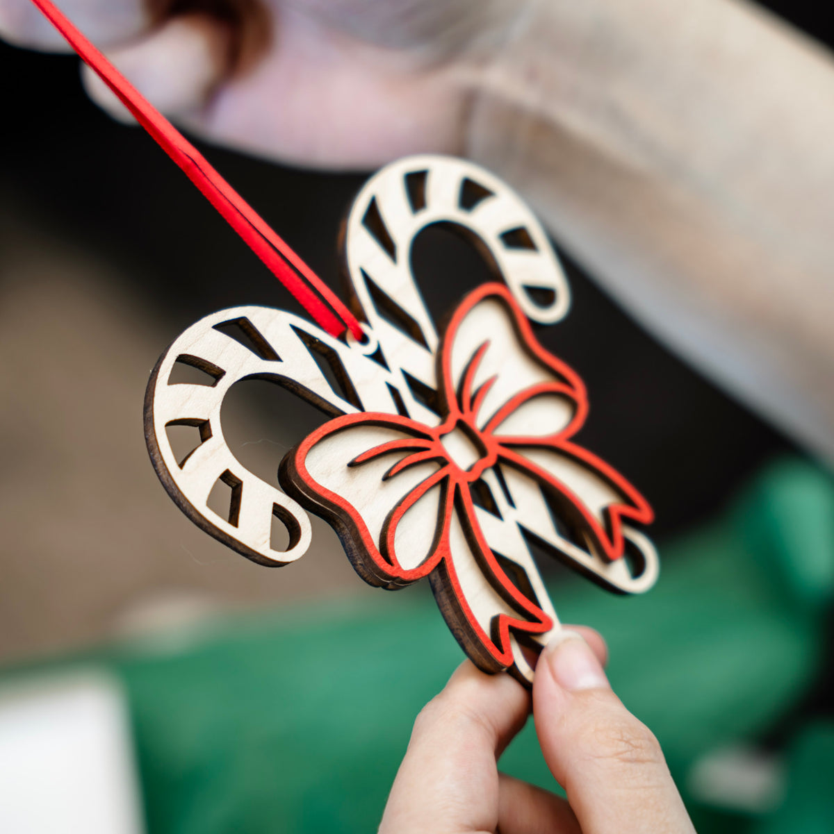 A close-up of hands holding a laser-cut wooden Christmas ornament shaped like two candy canes with a red bow, suspended by a red ribbon against a soft-focus background of green tissue paper and a gift box.