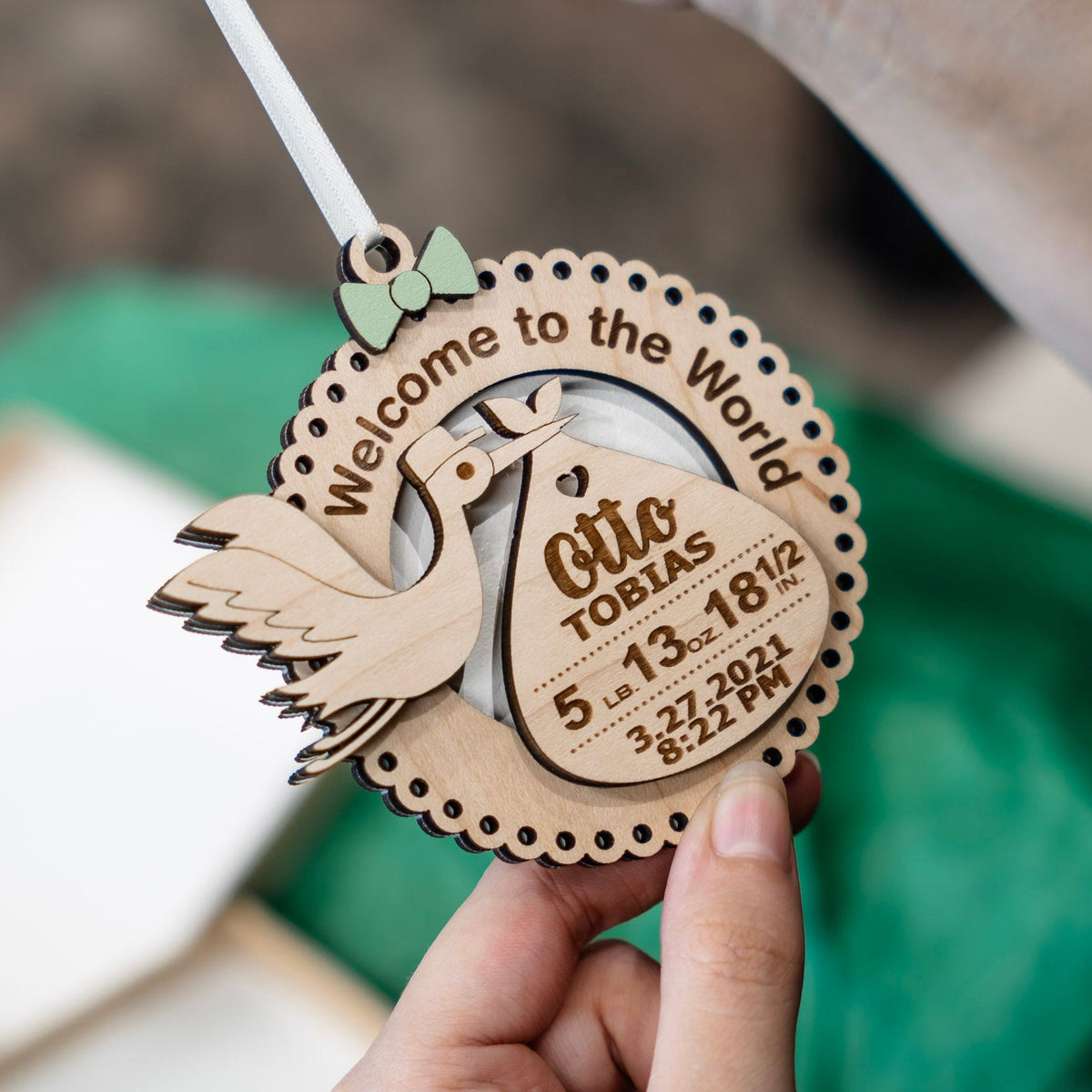 A hand holds a personalized wooden baby shower ornament shaped like a stork with a green bow, hung with a white ribbon, featuring engraved text reading 'Welcome to the World' and 'Otto Tobias' with birth details. The background is softly blurred with green fabric and neutral tones.