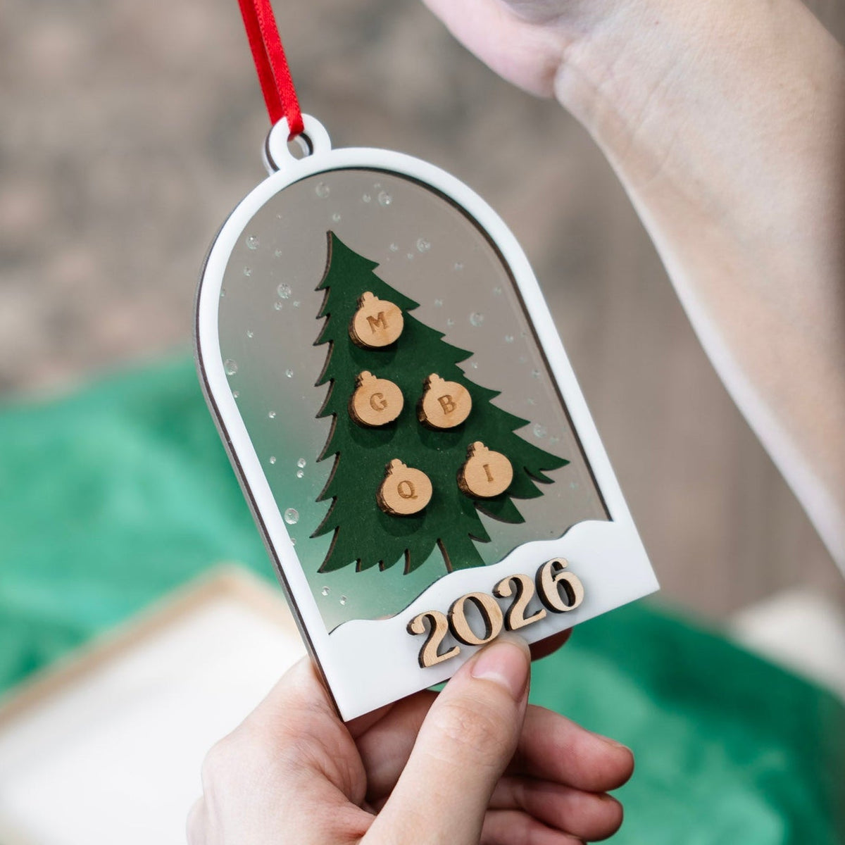 A person holds a personalized wooden Christmas ornament shaped like a tree with the year '2026' and small wooden ornaments for initials, hanging it by a red ribbon. The background is softly blurred with a green fabric surface.