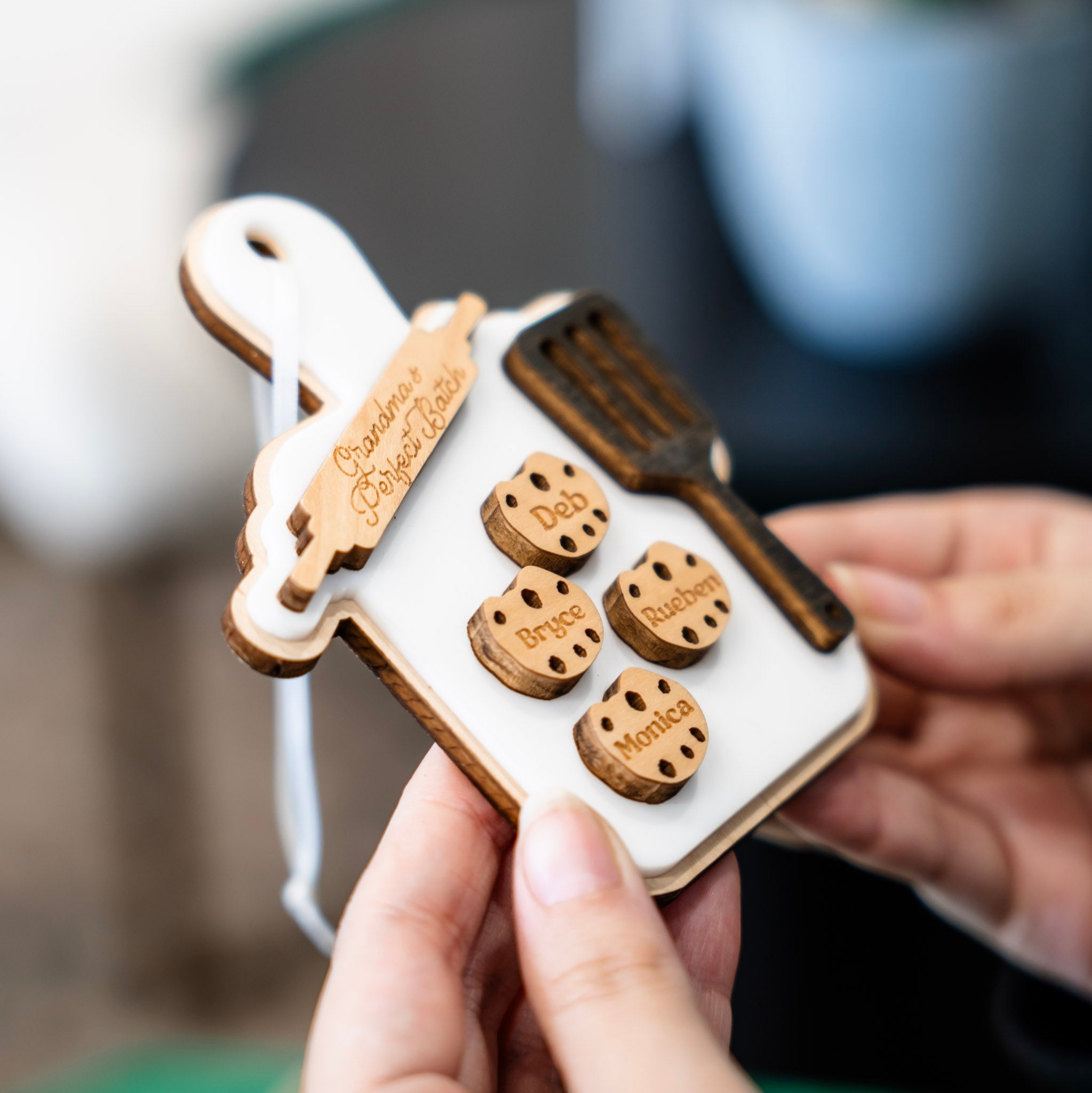 A personalized wooden baking-themed keepsake ornament held in two hands. The ornament is shaped like a white baking tray with a wooden rim, featuring a wooden rolling pin, a dark wooden spatula, and four round wooden cookie cutouts engraved with names: Deb, Bryce, Rueben, and Monica.