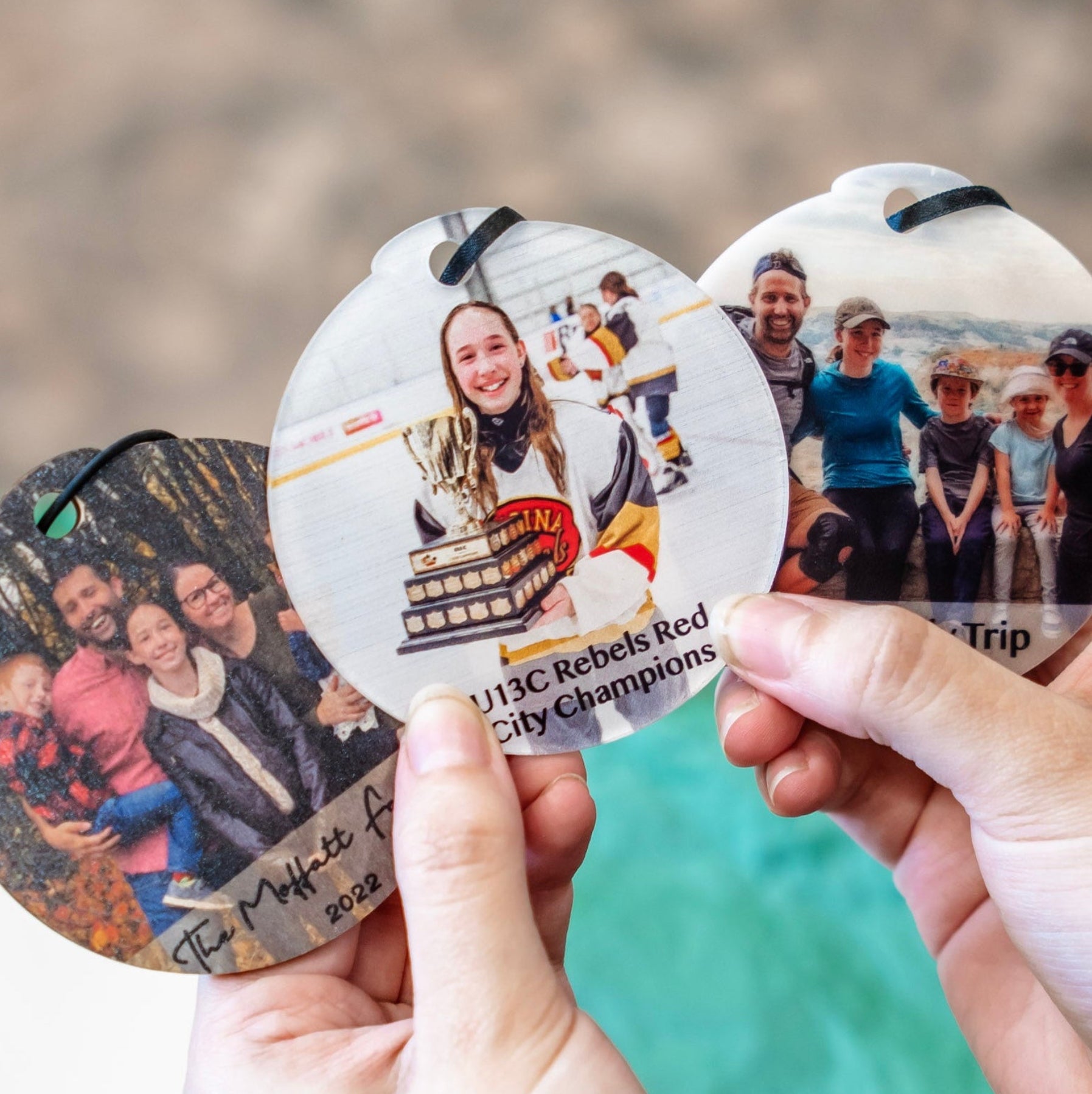 Three personalized round photo coasters held in two hands, showing custom printed family photographs with black hanging loops at the top.