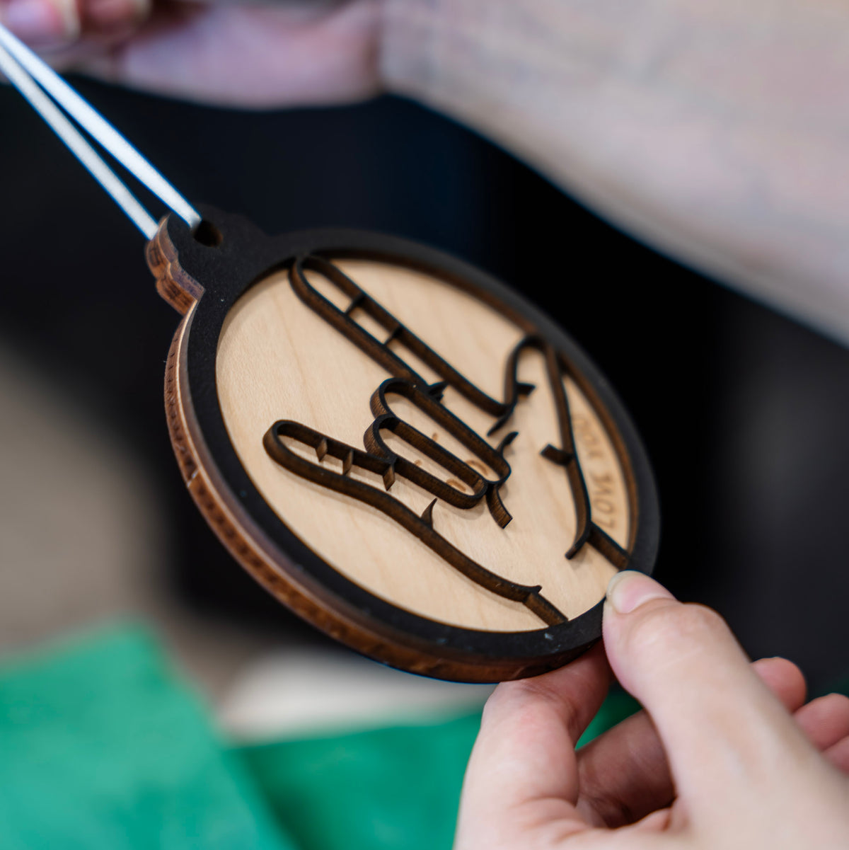 A person holds a round wooden ornament with a black border and a laser-cut hand design. The ornament has a light natural wood center with a black engraved hand signing  'I LOVE YOU' in ASL. 