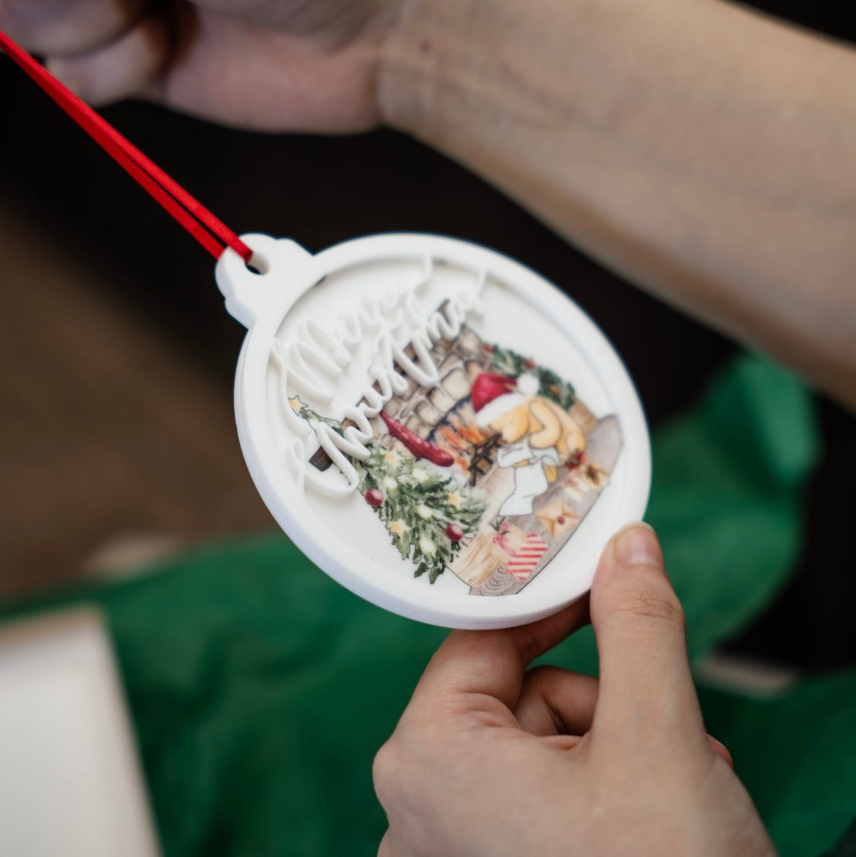A person holds a round white Christmas ornament with a red hanging string. The ornament features a layered cutout design with the words “Merry Christmas” over a festive Winnie the Pooh winter scene with a tree, fireplace, and gift boxes.