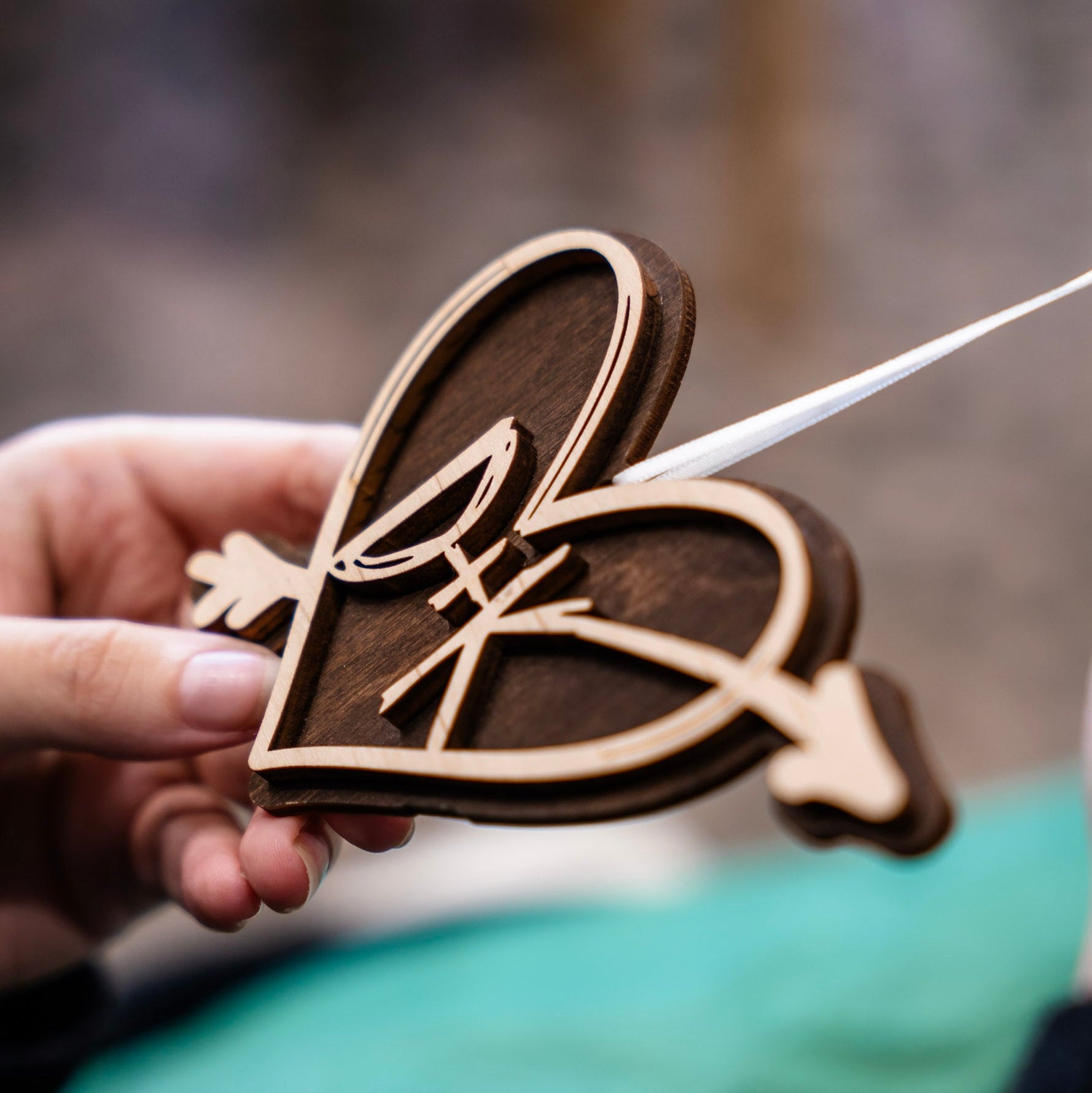 A close-up of a person holding a layered wooden heart-shaped ornament with an arrow through it and initials 'DK', showing natural wood tones and a white ribbon attached.
