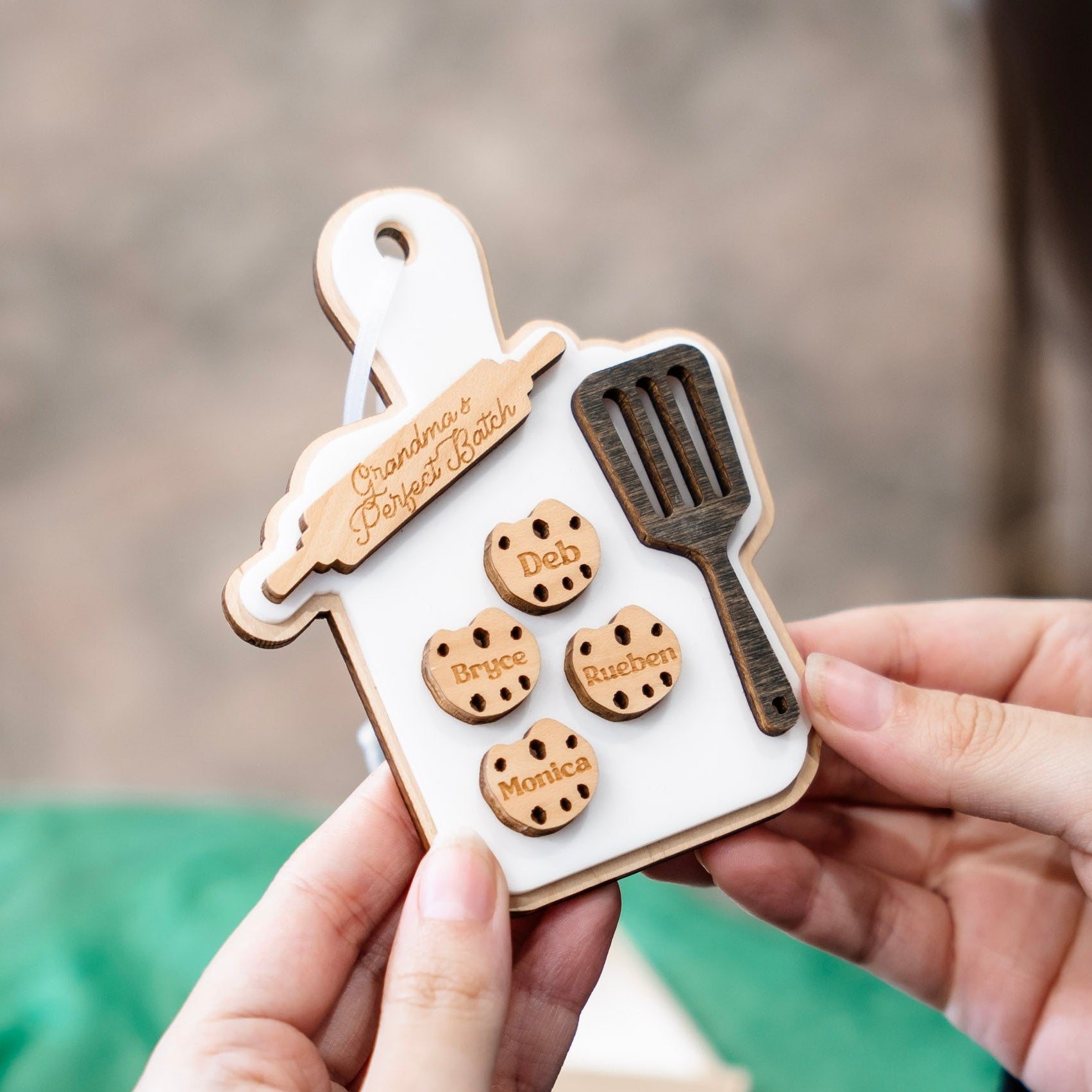 A personalized wooden baking-themed keepsake ornament held in two hands. The ornament is shaped like a white baking tray with a wooden rim, featuring a wooden rolling pin, a dark wooden spatula, and four round wooden cookie cutouts engraved with names: Deb, Bryce, Rueben, and Monica.
