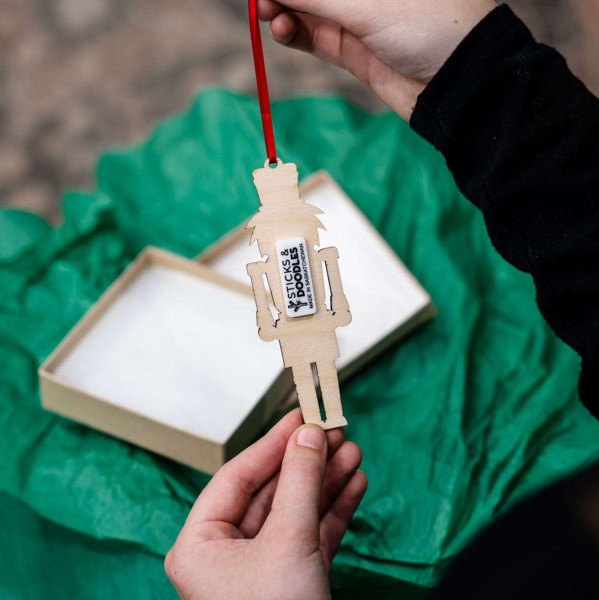 A person holds the back of a small wooden nutcracker ornament with a red hanging ribbon. The ornament has a white tag reading 'Sticks & Doodles made in Saskatchewan'.