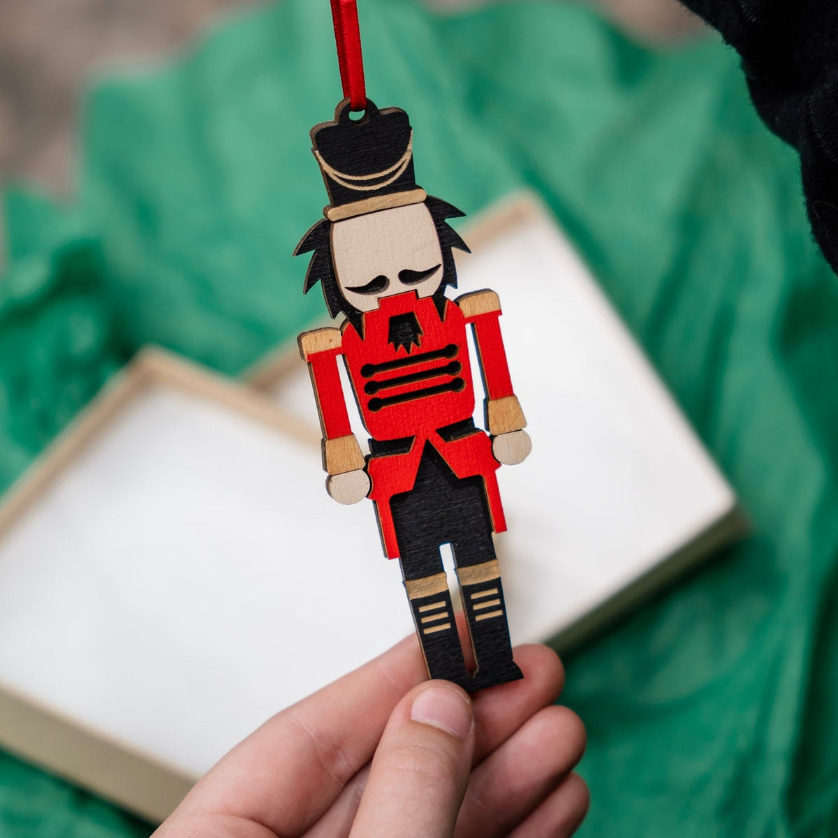 A hand holds a small wooden nutcracker ornament with a red hanging ribbon. There is green crumpled up tissue paper in the background behind a beige gift box.
