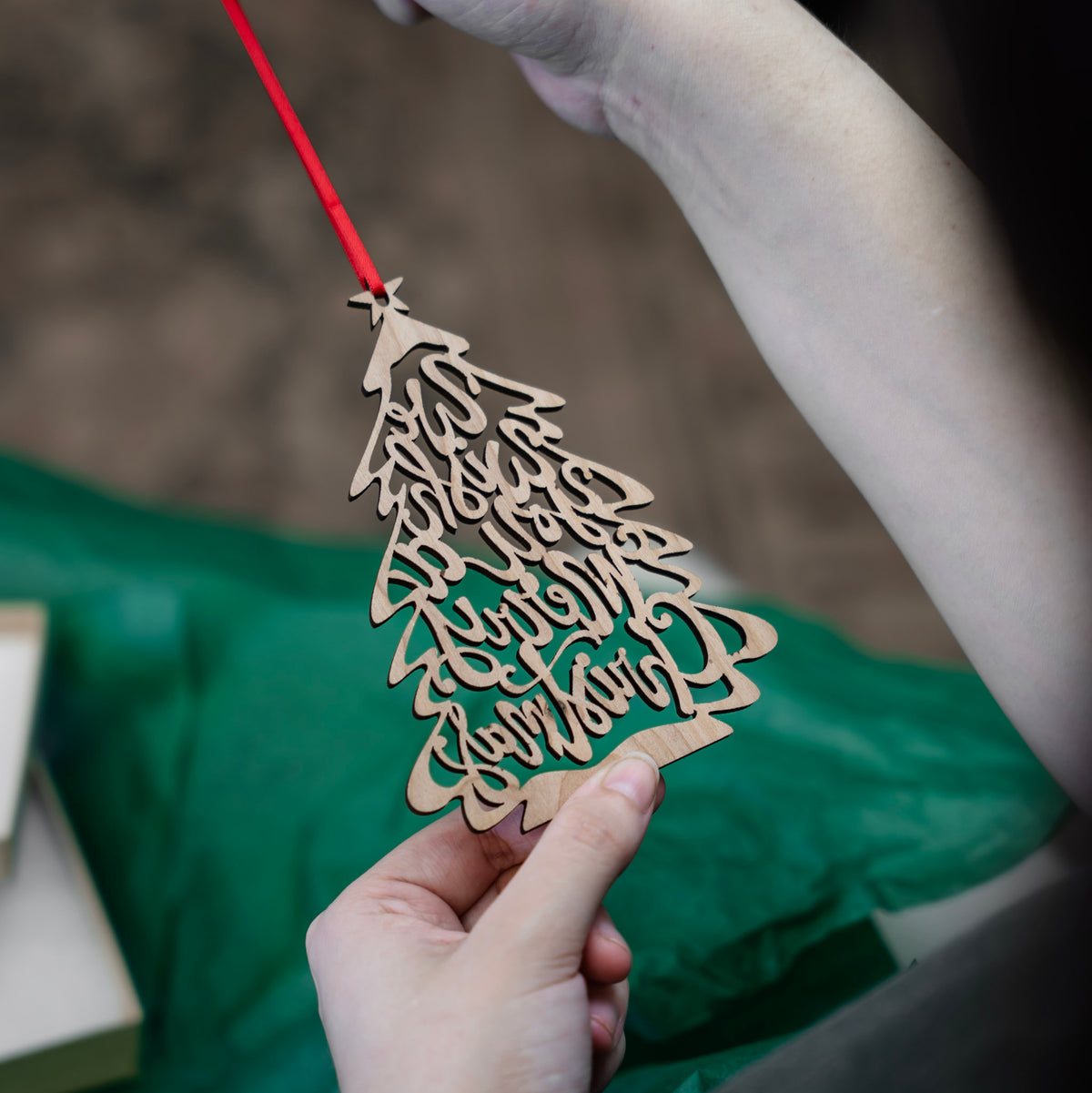 A close-up of a hand holding the back side of a laser-cut wooden Christmas tree ornament with 'We Wish you a Merry Christmas' written in the branches, suspended from a red ribbon. A person's other hand is holding it up against a soft-focus background with green tissue paper and gift box.
