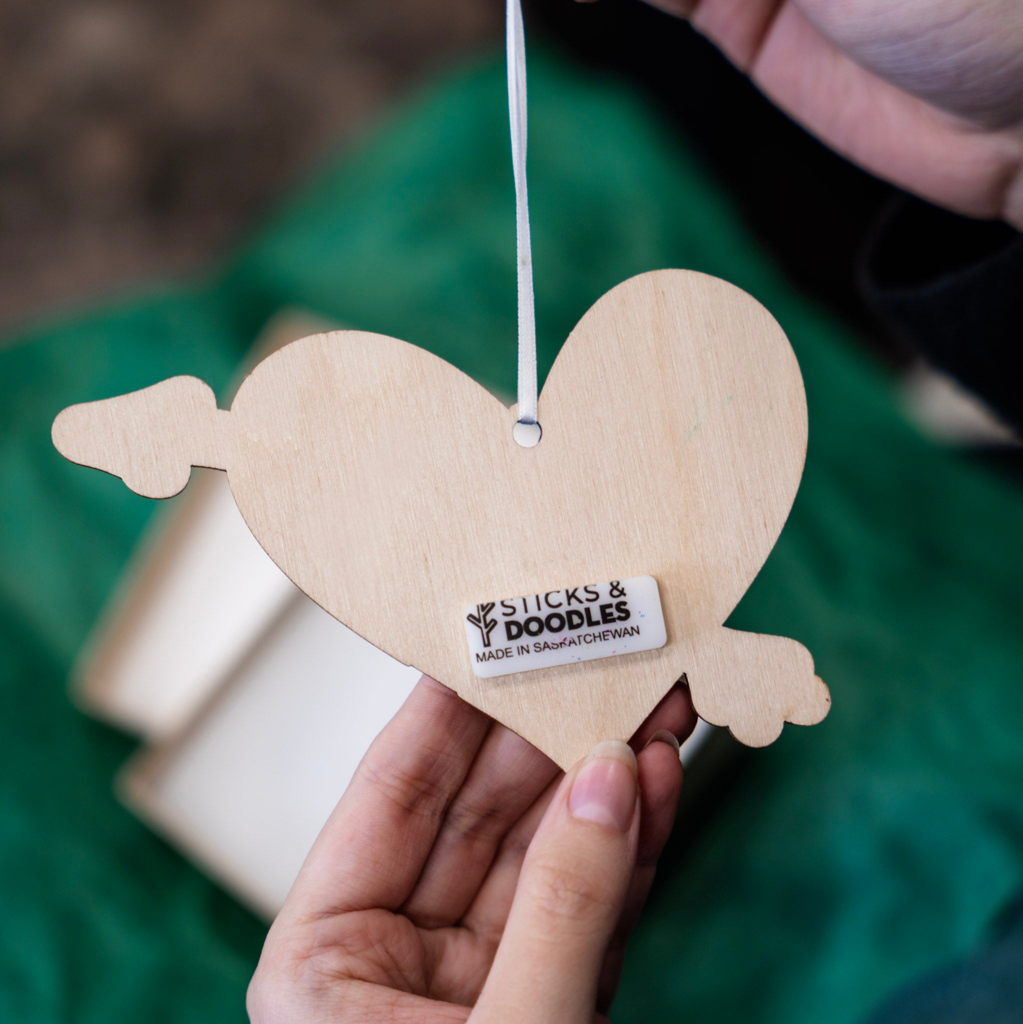 A hand holds a light natural wood heart-shaped ornament with an arrow cutting through, hanging with a white ribbon. A small label on the front reads 'STICKS & DOODLES MADE IN SASKATCHEWAN'.
