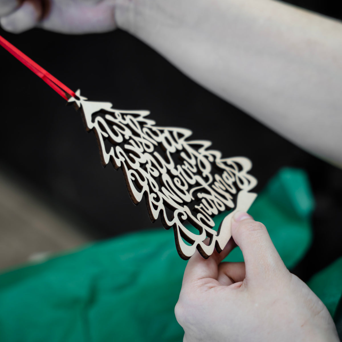 A close-up of a hand holding a laser-cut wooden Christmas tree ornament with 'We Wish you a Merry Christmas' written in the branches, suspended from a red ribbon. A person's other hand is holding it up against a soft-focus background with green tissue paper and gift box.