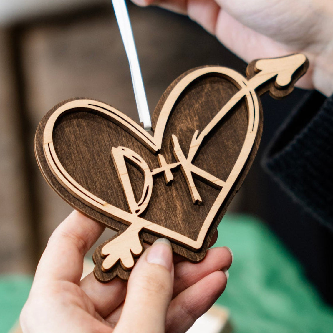 A person holds a wooden heart-shaped ornament with an arrow and the initials 'DK' in the center, suspended by a white ribbon. The background is softly blurred with a green fabric surface and neutral indoor setting.
