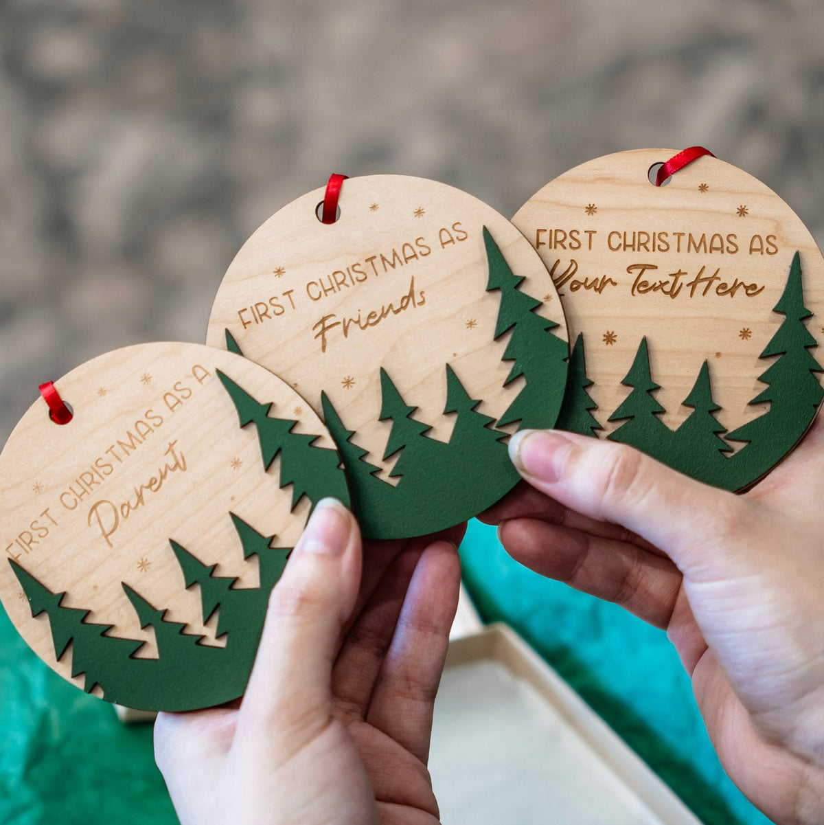 Three round wooden Christmas ornaments held in hands, featuring laser-engraved text options like 'First Christmas as a Parent', 'First Christmas as a Friend', and 'First Christmas as Your Text Here', with green pine tree silhouettes and red hanging loops.