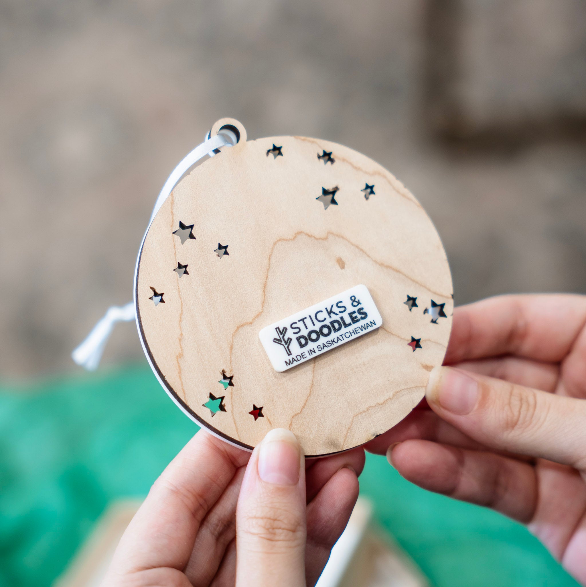A person holds a round wooden ornament with star-shaped cutouts and a white sticker reading 'STICKS & DOODLES MADE IN SASKATCHEWAN'.