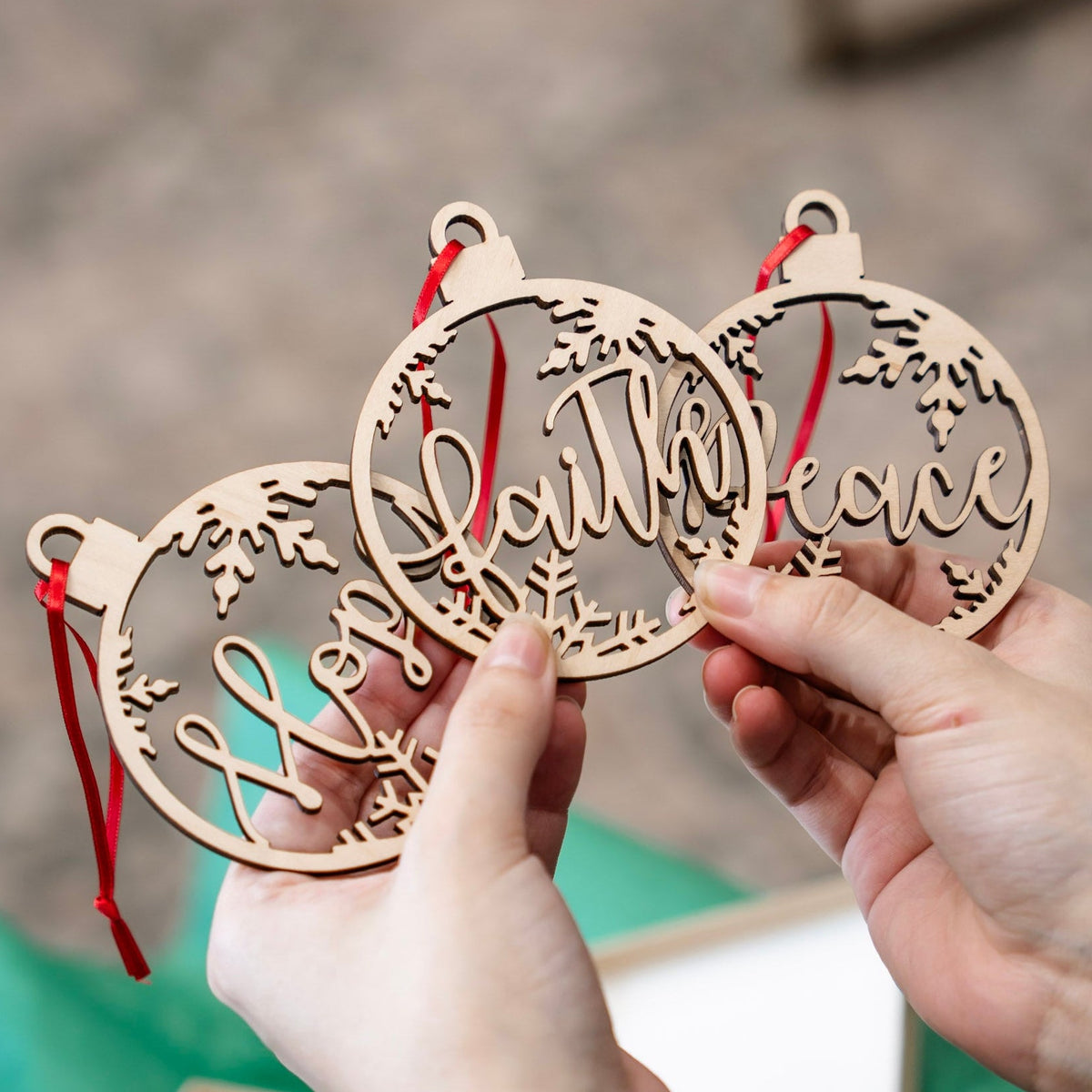 Three round wooden Christmas ornaments with red hanging ribbons are held in two hands. The ornaments are laser-cut with holiday designs and cursive words including 'Hope',  'Faith' and 'Peace'.
